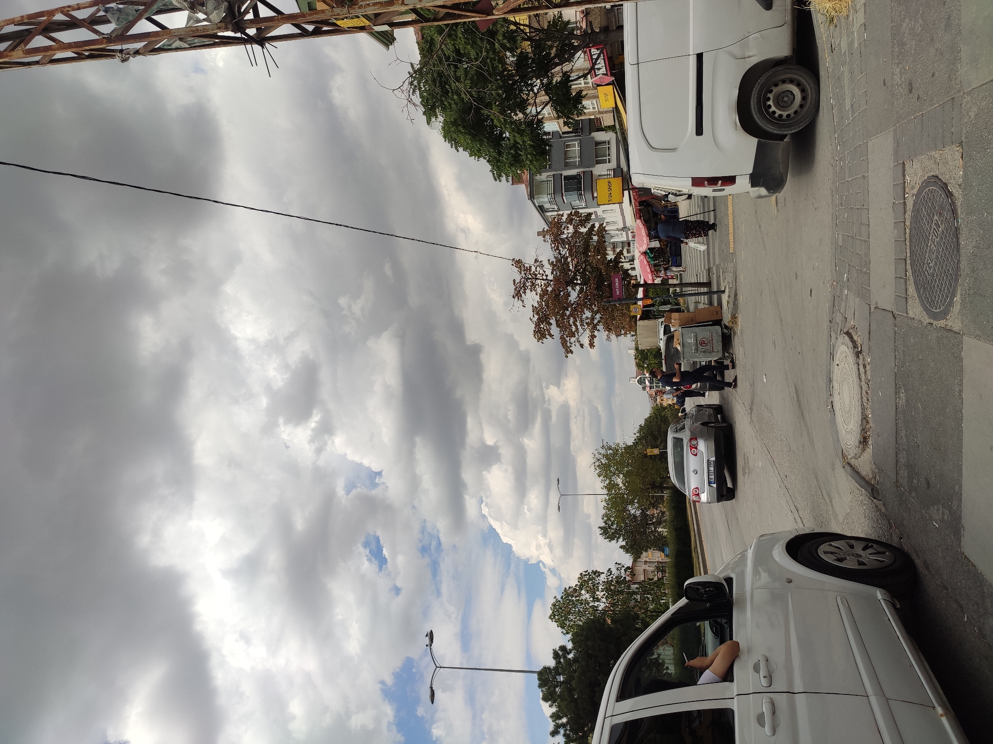 The image depicts a street scene in the Mamak district of Ankara, Turkey, under a cloudy sky. A white van is parked on the right side of the frame, while another white van is partially visible on the left, with a person's arm resting on its open window. A silver sedan is driving on the road in the middle ground. A man is standing near the silver sedan, and a woman with a headscarf and a white bag is walking on the sidewalk. Other pedestrians and market stalls are visible further down the street. A utility pole with wires stands on the right. The environment includes buildings, trees, streetlights, and a trash bin.