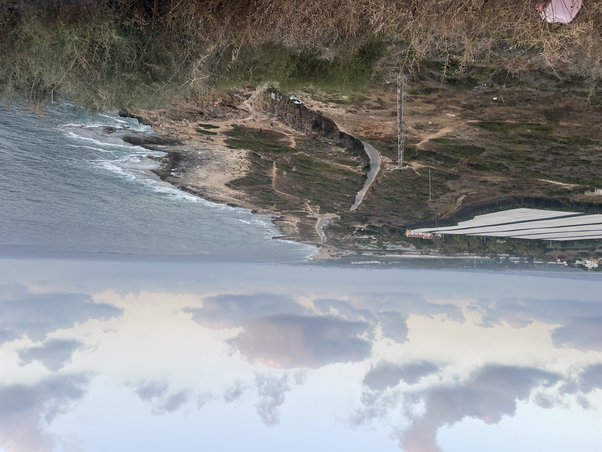 The image depicts a coastal landscape in the Mate Asher Regional Council, Israel, under a cloudy sky. In the foreground, dry brush and vegetation are visible. A dirt path winds down a hillside towards a rocky coastline where the sea meets the shore with gentle waves. A narrow road follows the coastline, with several vehicles parked along it. Further inland, a large agricultural greenhouse complex is situated on a hillside. A tall metal utility pole stands on the slope. In the distance, buildings and a town are visible along the coast. A white car is parked on a dirt path near a rocky outcrop overlooking the sea.