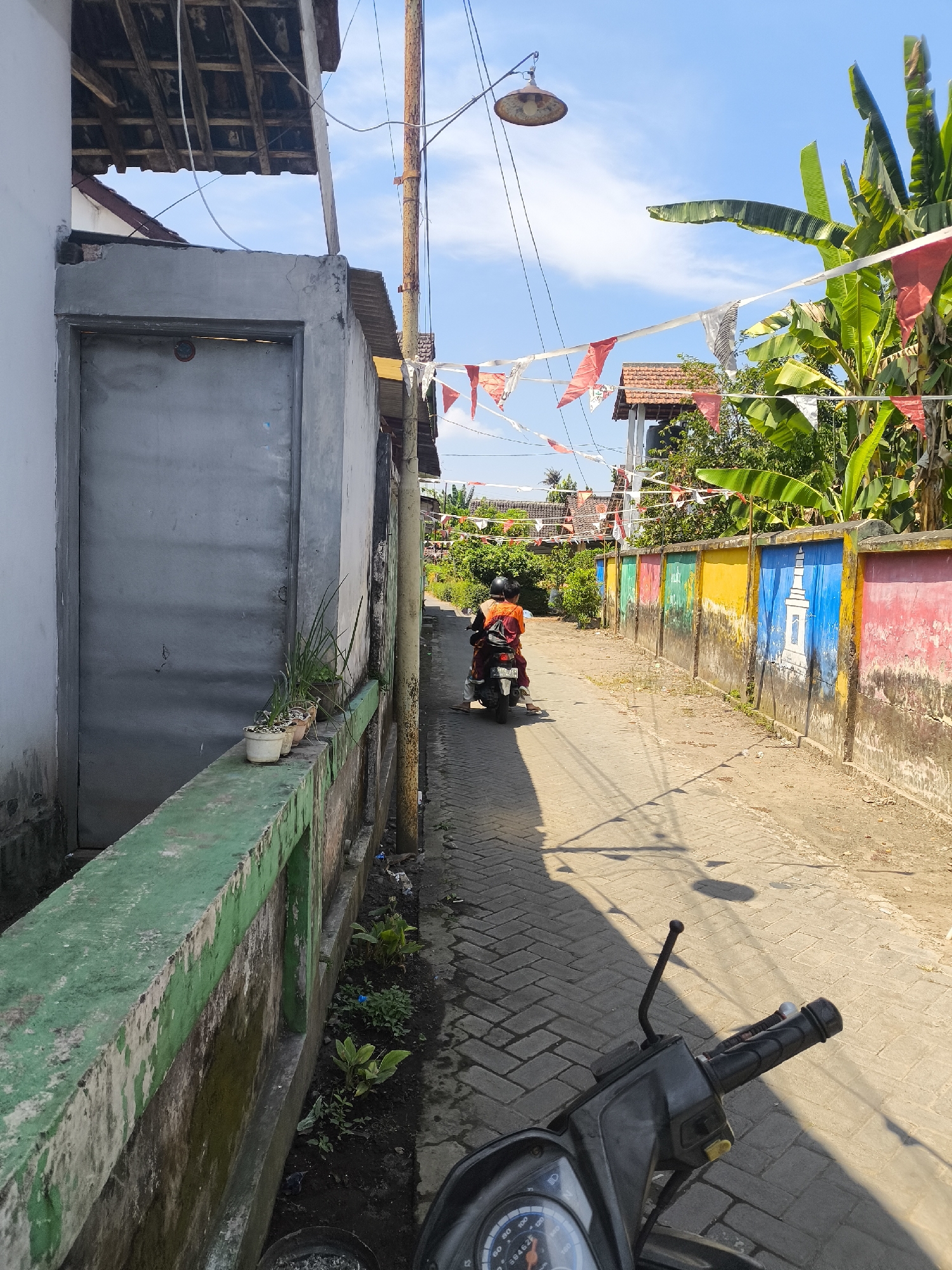 In Lumajang, Indonesia, a narrow, paved alleyway runs between buildings on a sunny day. A person on a motorcycle, wearing an orange shirt and helmet, is traveling down the alley away from the camera. The alley is lined with a colorful mural on the right side of the wall, featuring various colors and designs, including a depiction of a white monument. Small red and white flags are strung across the alley overhead, suggesting a celebration or national holiday. On the left side of the alley, a gray wall with a metal door is visible, and a low green-painted wall runs along its base, with potted plants on top. The ground is paved with interlocking bricks, and shadows from the overhead structures and trees are cast across the surface. Lush green foliage, including banana trees, is visible in the background. The overall scene depicts a residential area in Lumajang.