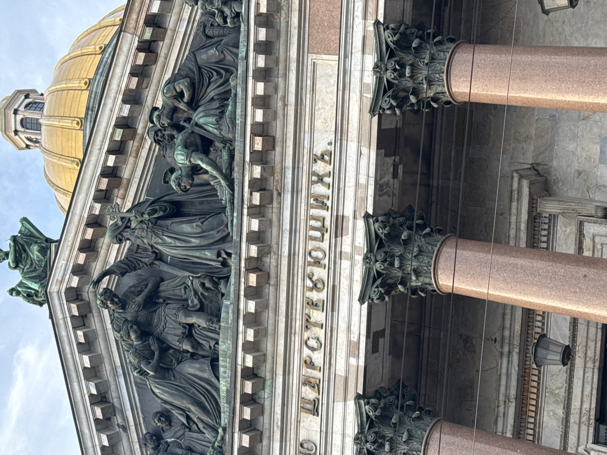 The image provides an upward-angled view of a grand architectural edifice in Saint Petersburg, Russia, unmistakably St. Isaac's Cathedral. A segment of its iconic golden dome is visible against a bright, partly cloudy sky. The building's facade is adorned with massive classical columns, likely made of red granite, topped with ornate bronze Corinthian capitals.
Beneath the dome and above the columns, a prominent high-relief sculptural frieze extends across the facade. This intricate bronze artwork depicts numerous figures in various states and poses, suggesting a dramatic historical or religious narrative. The figures are clad in draped garments; some appear in distress or supplication, while others adopt more stoic or commanding postures. One figure is seen holding a cross, and others resemble soldiers or civic leaders, creating a complex and emotive tableau. Decorative lantern fixtures are visible, hanging from the structure.
The photograph captures the scene during daylight hours, indicated by the clear illumination. The overall impression is one of monumental scale and rich historical detail.
Gilded Cyrillic lettering is prominently displayed on the facade, integrated into the architectural design. The visible text reads "ЦАРСТВУЮЩИХЪ ДОБРОДѢТЕЛИ", which translates to "VIRTUES OF RULERS" or "THE REIGNING VIRTUES," forming part of a larger inscription found on the cathedral's pediments.