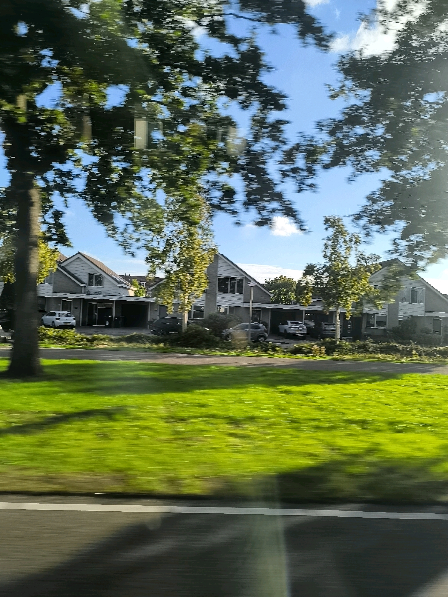 This is a side-view, motion-blurred image taken from a vehicle, likely in the city of Oostvoorne, Netherlands, on a sunny day with a clear blue sky dotted with clouds. The scene is dominated by a residential area featuring modern, two-story houses with grey roofs and light-colored facades. Many of the houses have attached carports with vehicles parked underneath. A bright green, well-maintained lawn stretches across the foreground, bordering a paved road. Mature trees with green foliage are prominent throughout the scene, their leaves creating a dappled effect and contributing to the motion blur. In the background, more trees and the sky are visible. The overall impression is that of a suburban neighborhood captured during a drive.