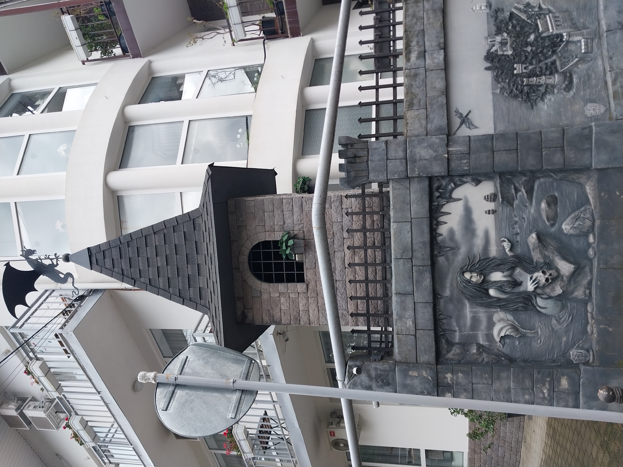 The image shows a whimsical building structure in Sochi, Russia, featuring a turret-like extension with a pointed roof and a weather vane shaped like a dragon. The structure is adjacent to a modern building with white facade and curved architectural elements. A decorative wall with bas-reliefs depicting mythical scenes, including a mermaid-like figure, is incorporated into the lower portion of the structure. A street sign is visible in the foreground.