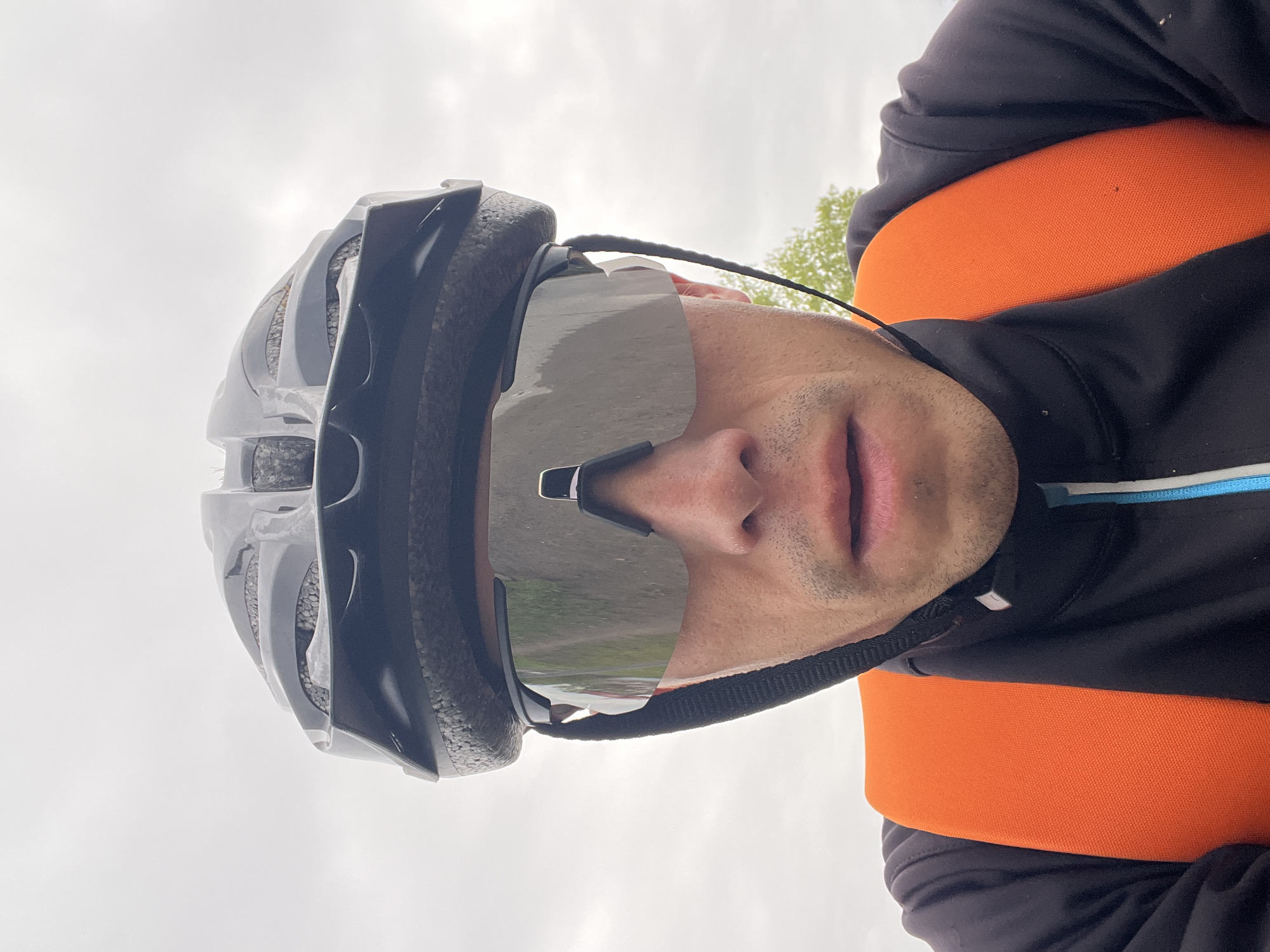 A person is wearing a black cycling helmet with clear cycling glasses, a black jacket with a blue and white stripe on the shoulder, and an orange strap around the waist. The person is likely outdoors, as a cloudy sky is visible above, and a blurred landscape appears reflected in the glasses. The location is unspecified, and there is no definitive evidence to place this image in Chaikovsky, Russia.