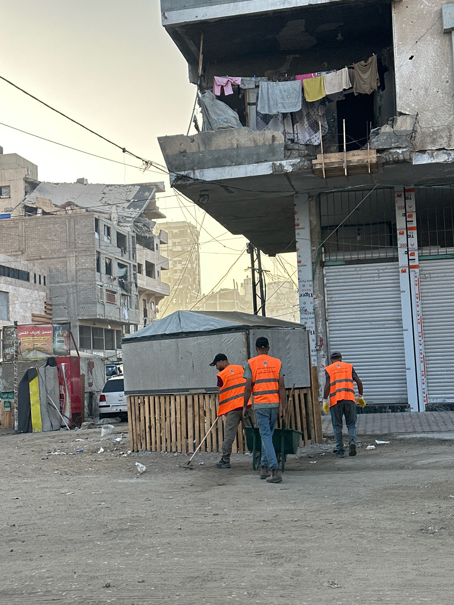 In the city of Gaza, Palestinian Territories, three individuals are seen wearing bright orange safety vests, indicating they are likely involved in some form of cleanup or restoration work. The environment appears to be a street or open area, with debris and some building structures in the background. One individual in the foreground is actively sweeping the ground with a broom. Behind him, two other individuals are walking, one pushing a green wheelbarrow filled with tools. The buildings in the background show signs of damage, with some appearing partially destroyed or under construction, suggesting a post-conflict or ongoing development setting. The overall scene conveys a sense of ongoing efforts to rebuild or maintain order in a challenging urban environment.