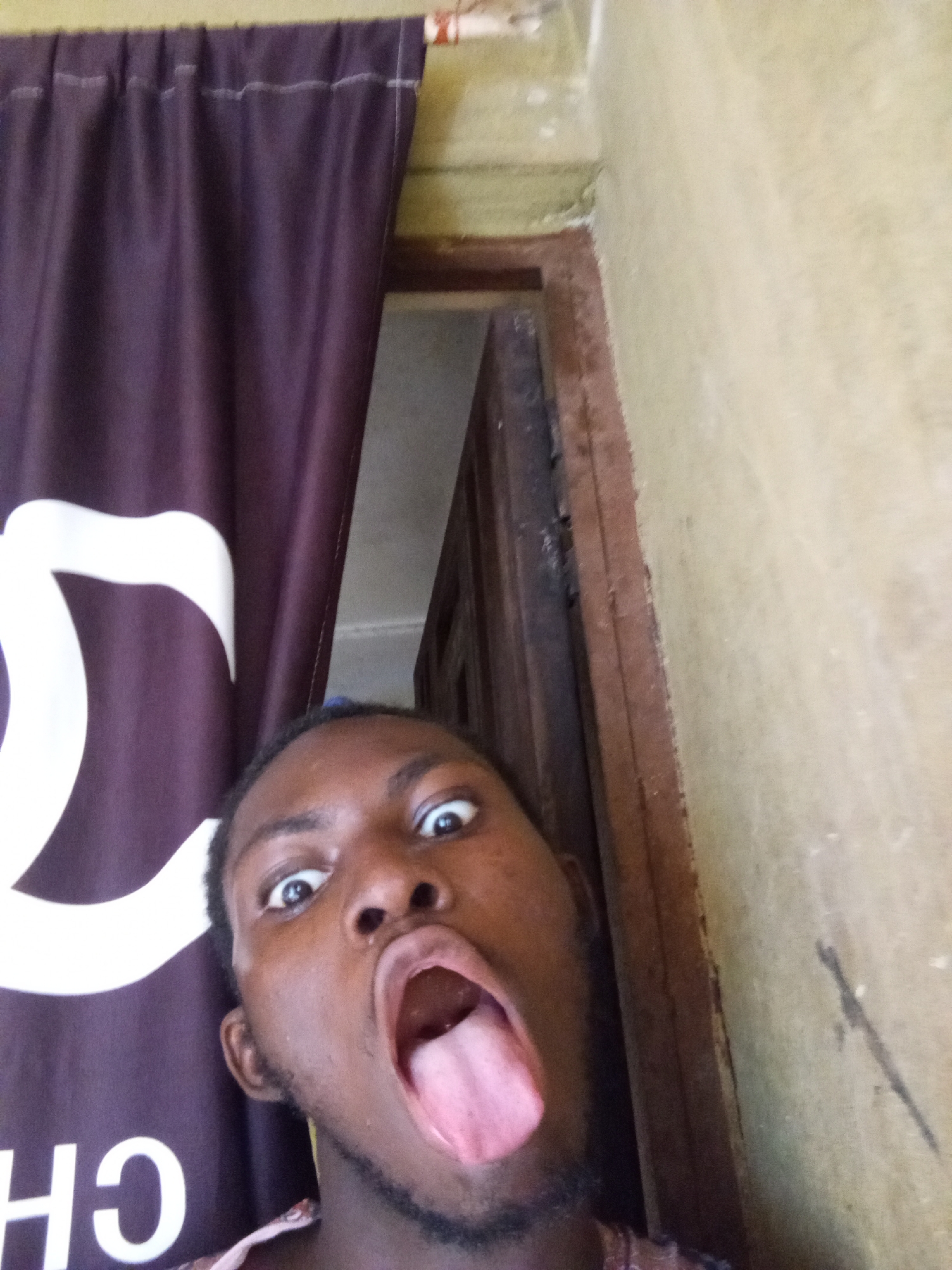 A selfie taken by a dark-skinned individual with their mouth open and tongue extended. The person has wide eyes and a dark beard. Behind the person is a dark-colored curtain with white lettering, and a wooden doorway. The location appears to be indoors with painted walls. The city and location are unknown.