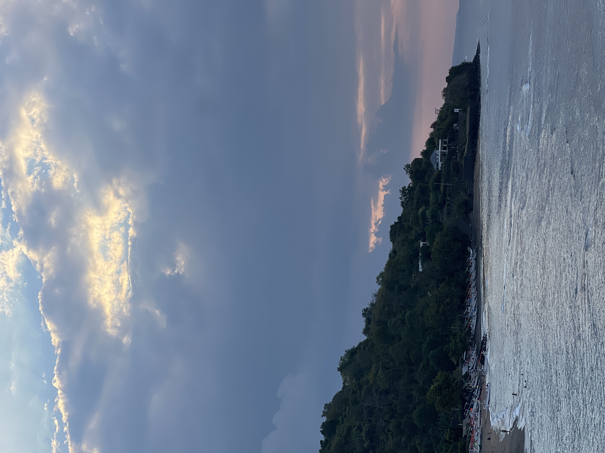 The image depicts a twilight scene on a beach in Abang, Indonesia. The sky is filled with dramatic clouds, with rays of sunlight breaking through, illuminating the upper portion of the frame with golden light. The lower part of the sky is a gradient of muted blues and soft pinks, indicating the transition from day to night.
A lush, green, tree-covered hillside slopes down to the beach. Along the shoreline, numerous traditional Indonesian outrigger boats are lined up on the dark sand. Several people are visible on the beach and in the shallow water, some wading and others possibly swimming. The ocean itself is calm, with gentle waves lapping the shore. A few buildings are nestled on the elevated terrain along the coastline. The overall atmosphere is serene and tranquil.