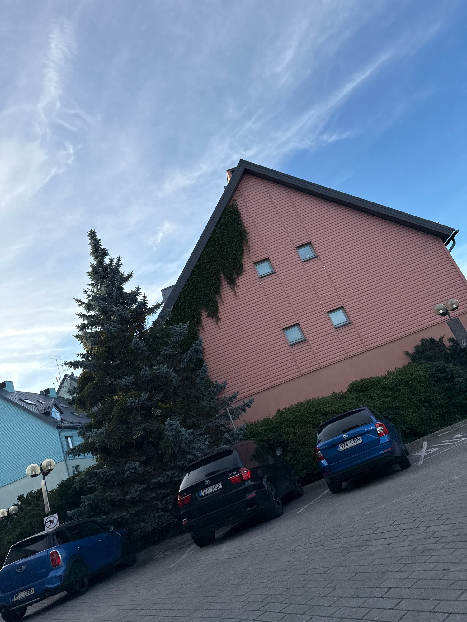 The image depicts a street scene in Tallinn, Estonia. In the foreground, a blue MINI Cooper is parked, followed by a black BMW X5. To the right, a blue Skoda Octavia is parked. The cars are situated in a paved parking area. Behind the cars, a large evergreen tree stands next to a pink building with a dark roof. The building has several small, square windows. To the left of the tree, the corner of a light blue building is visible. The sky above is blue with scattered white clouds. A street lamp with two globes is present on the right side of the image, near the pink building.