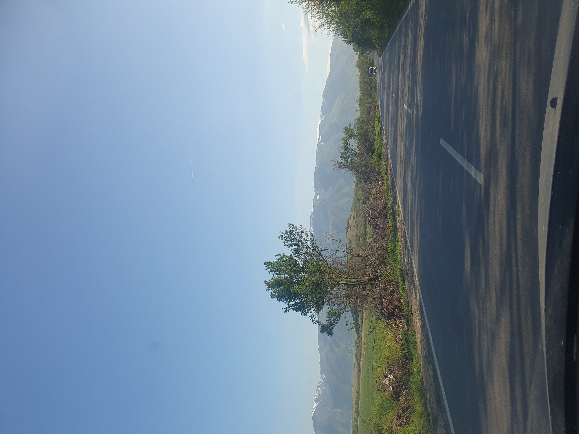 The image, when correctly oriented, captures a scenic roadside view through a mountainous region, specifically Marino Pole, Bulgaria. An asphalt road dominates the right side of the frame, extending into the distance. It features clear white dashed lines marking the lanes and a solid white line along its right edge. Long, dark shadows, likely cast by unseen trees, stretch across the road, suggesting early morning or late afternoon sunlight.
To the left of the road, a lush green landscape unfolds, starting with dense bushes and a prominent leafy tree in the foreground. Beyond this, a wide expanse of green fields leads to a series of distant, hazy mountains. Several of these mountains display distinct patches of white, indicative of snow or bright rock faces.
The sky, occupying the upper left quadrant, is a clear, bright blue with minimal cloud cover, suggesting fair weather. A faint, thin white contrail from an airplane is visible high in the sky. Far down the road, a small, light-colored vehicle, possibly a car or van, can be seen, highlighting the vastness of the road ahead. The scene conveys a sense of peaceful travel through a beautiful natural environment. No text is visible in the image.