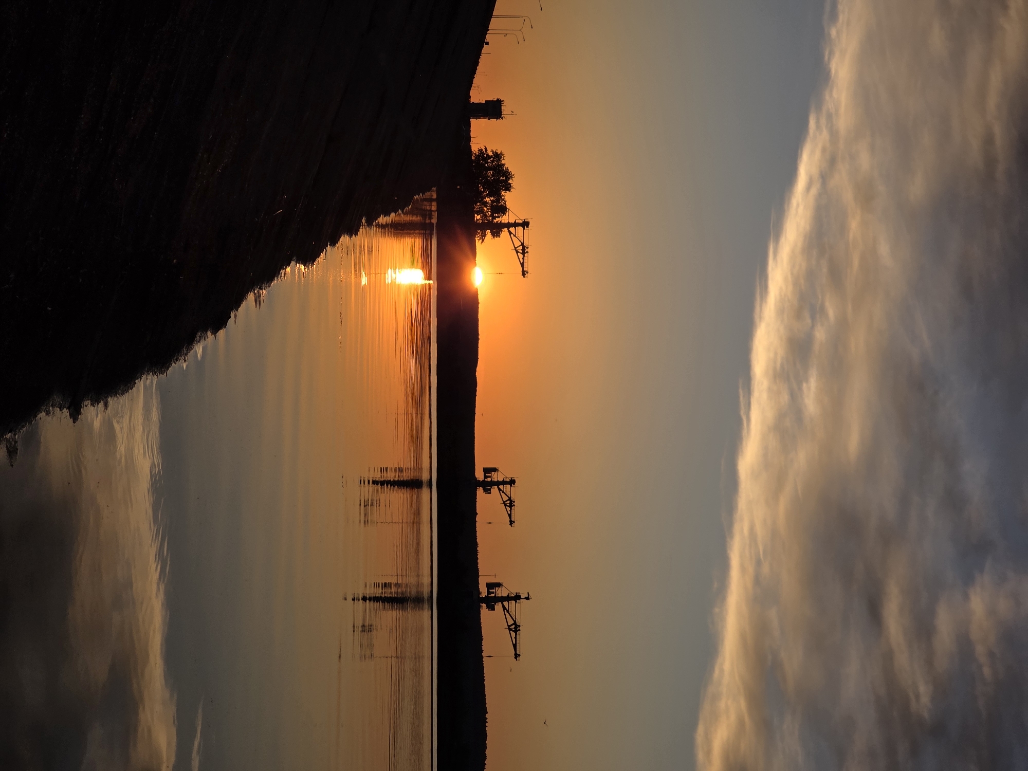 This image captures a vibrant sunset over a wide body of water, likely the Kama River near Chaikovsky, Russia. The sun, a brilliant orb of orange light, is positioned low on the horizon, creating a striking silhouette against the sky. Its intense radiance casts a shimmering, golden reflection that stretches across the calm surface of the water towards the foreground.
The distant shoreline is presented as a dark, irregular silhouette, indicating a landmass adorned with trees and sparse vegetation. Along this shadowed horizon, several distinct industrial structures, appearing as large cranes or excavators, stand tall, their mechanical forms sharply outlined against the fiery sky. These structures, along with the sun and shoreline, are clearly reflected in the tranquil water below, though with some subtle distortion from gentle ripples.
The sky above the horizon displays a beautiful gradient of colors, transitioning from intense oranges and reds immediately surrounding the sun to softer peach and muted blue tones further upwards. To the right, a large, voluminous cloud formation is illuminated by the setting sun, glowing with golden and warm hues against the sky, and its detailed shape is also mirrored in the water.
The scene conveys a serene and peaceful atmosphere, characteristic of late evening. The dramatic interplay of light and shadow, combined with the industrial elements along the natural landscape, creates a compelling visual. There is no visible text in the image.