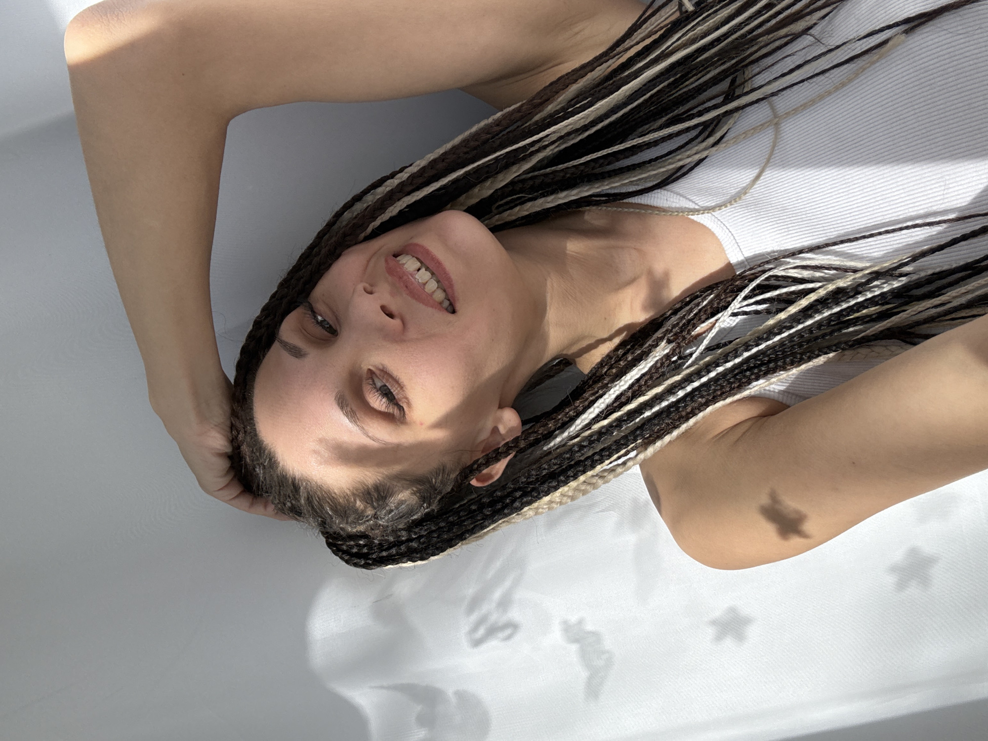 This close-up portrait, captured in Rusanovo, Russia, features a woman lying down indoors, looking directly up at the camera with a relaxed, smiling expression. Her multi-toned braids, a mix of black, brown, and blonde strands, are spread around her head and shoulders. She is wearing a simple white ribbed tank top. Her arms are bent and raised, with her hands resting near her head. The background consists of a plain, light-colored surface, possibly a floor or wall, suggesting a minimalistic indoor setting. Bright natural light illuminates the scene, creating sharp contrasts and casting distinct, decorative star-shaped shadows onto the surface in the lower right area of the image. The time of day appears to be daytime, indicated by the strong, direct sunlight. The woman's open-mouthed smile, revealing her teeth, conveys a cheerful and content emotion. There is no visible text in the image.