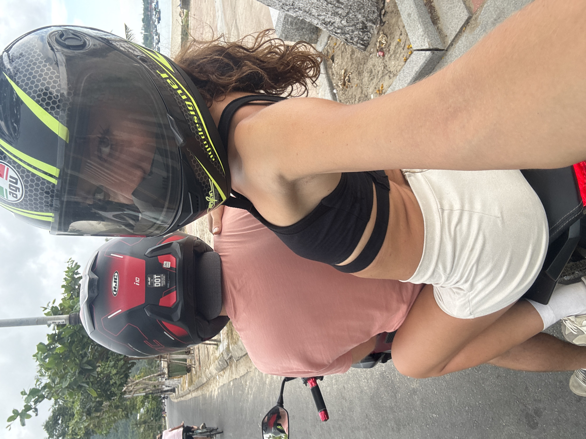 A woman wearing a black crop top and white shorts is riding on the back of a motorcycle being operated by a person wearing a pink shirt. Both people are wearing helmets. The woman is taking a selfie. The environment appears to be a road in a coastal urban area. The location is likely in the city of Đà Nẵng, Vietnam, based on the visual clues present.