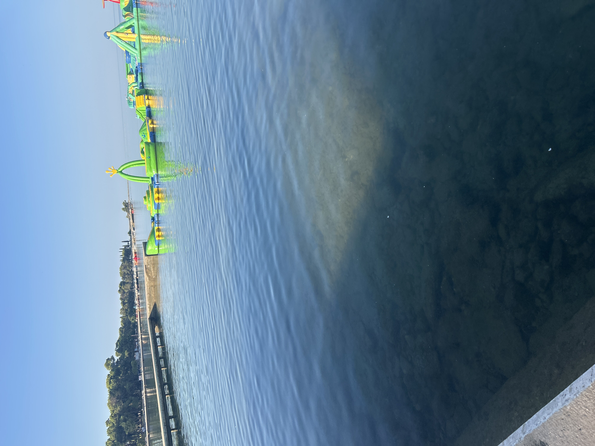 The image captures a tranquil daytime scene at the water's edge in Katoro, Croatia. In the foreground, clear blue water exhibits ripples, revealing the rocky seabed beneath. To the left, a concrete pier extends into the water, its surface textured. Dominating the middle ground and extending into the background is a vibrant inflatable water park, featuring various structures like slides, climbing walls, and obstacle courses in shades of green and yellow. The water park floats on the calm sea, reflecting its bright colors. Beyond the water park, a treeline of lush green trees marks the shoreline, with scattered buildings and possibly beach umbrellas visible amongst them. The sky above is a clear, cloudless blue, suggesting fair weather. No people are clearly visible within the frame.