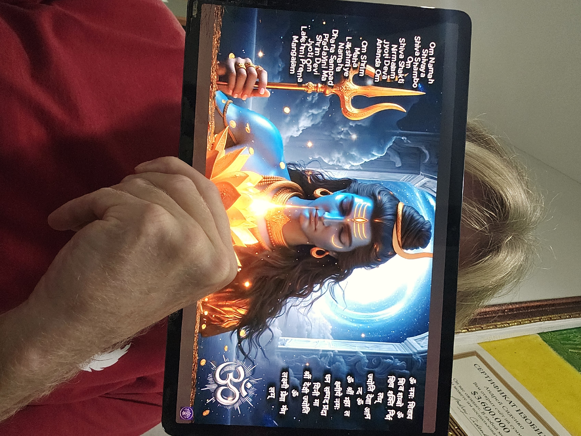 An indoor shot features a person, likely male, with fair skin and light hair, holding a black tablet. The tablet is angled, displaying a vibrant image of Lord Shiva with closed eyes, a glowing aura, and holding a golden trishul against a celestial background. The person's left hand is visible, holding the tablet from the side, while part of another person's light-colored hair is visible on the right side of the frame.
The tablet screen prominently features Hindu mantras written in both English transliteration and Devanagari script. The English text includes "Om Namah Shivaya," "Shiva Shambo," "Shiva Shakti Nirmalam," "Jyoti Deva Ananda Om," "Om Shrim Maha Lakshmiye Namaha," "Dhana Sampad Pradayini Ma," "Shrim Devi Jyoti Om," and "Lakshmi Prema Mangalam." A large "ॐ" symbol is also visible on the screen.
In the background, to the right of the tablet, a partially visible certificate with Russian text is seen. The certificate is titled "СЕРТИФИКАТ ИЗОБИЛИЯ" (Certificate of Abundance) and mentions "Андрей Сиденко" with an amount of "$3,690.000." The text further states, "Вам открыт счёт на сумму $3,690.000. Он вас ожидает. В любой момент вы можете им воспользоваться" (An account has been opened for you for the sum of $3,690.000. It awaits you. At any moment you can use it), and is signed "Andrej Cident". The scene suggests a personal setting, possibly in Denpasar, Indonesia, where spiritual content is being viewed.