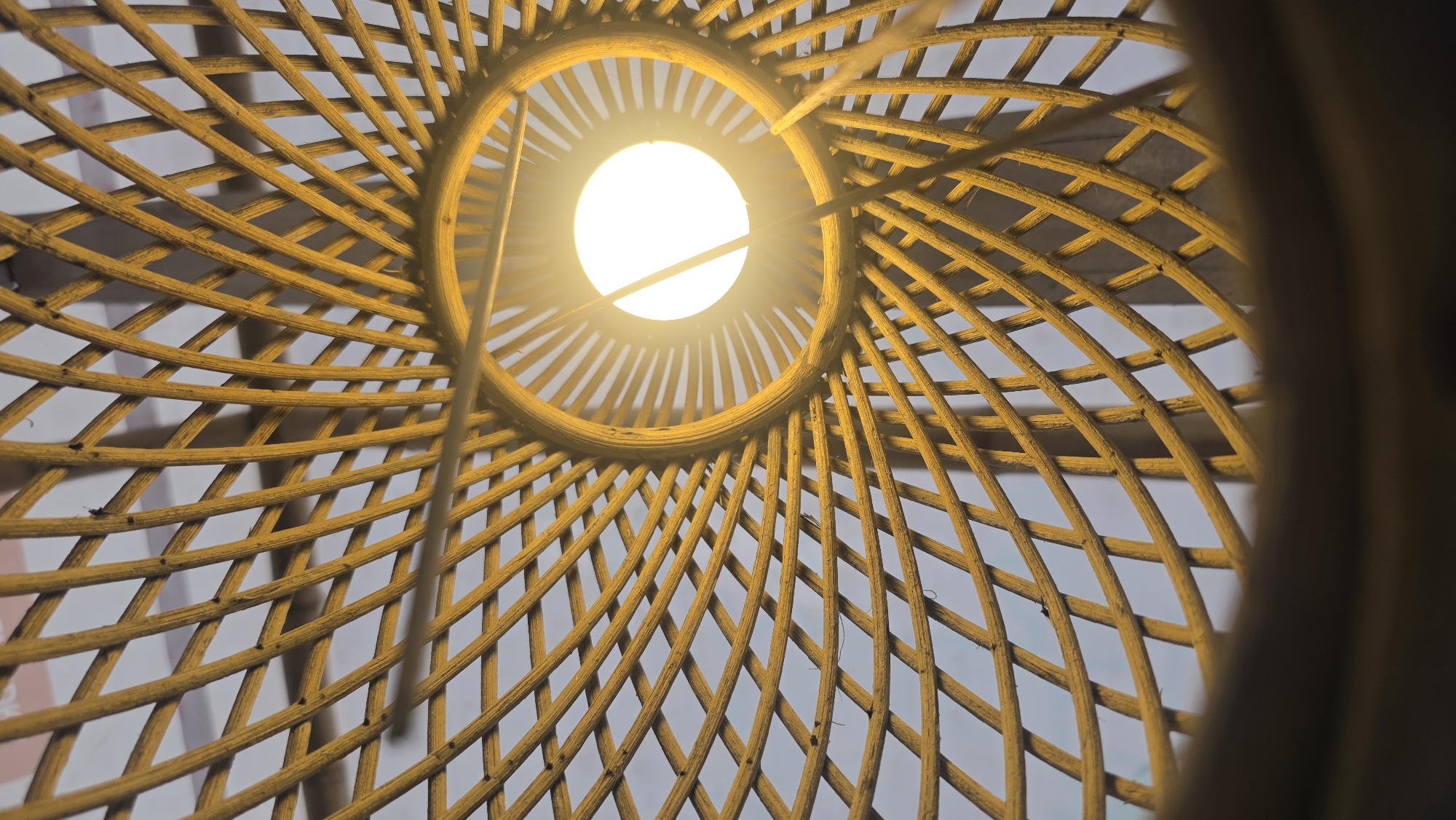 The image is a close-up, upward view of a woven, spherical lampshade with a lit bulb inside. The shade is constructed from thin, curved strands of a light-colored material, possibly bamboo or rattan, interwoven to create a mesh pattern. The light bulb at the center appears bright and white. The scene is presumably indoors. The image does not provide enough information to confirm the location in the city of Kuala Pidie, Indonesia.