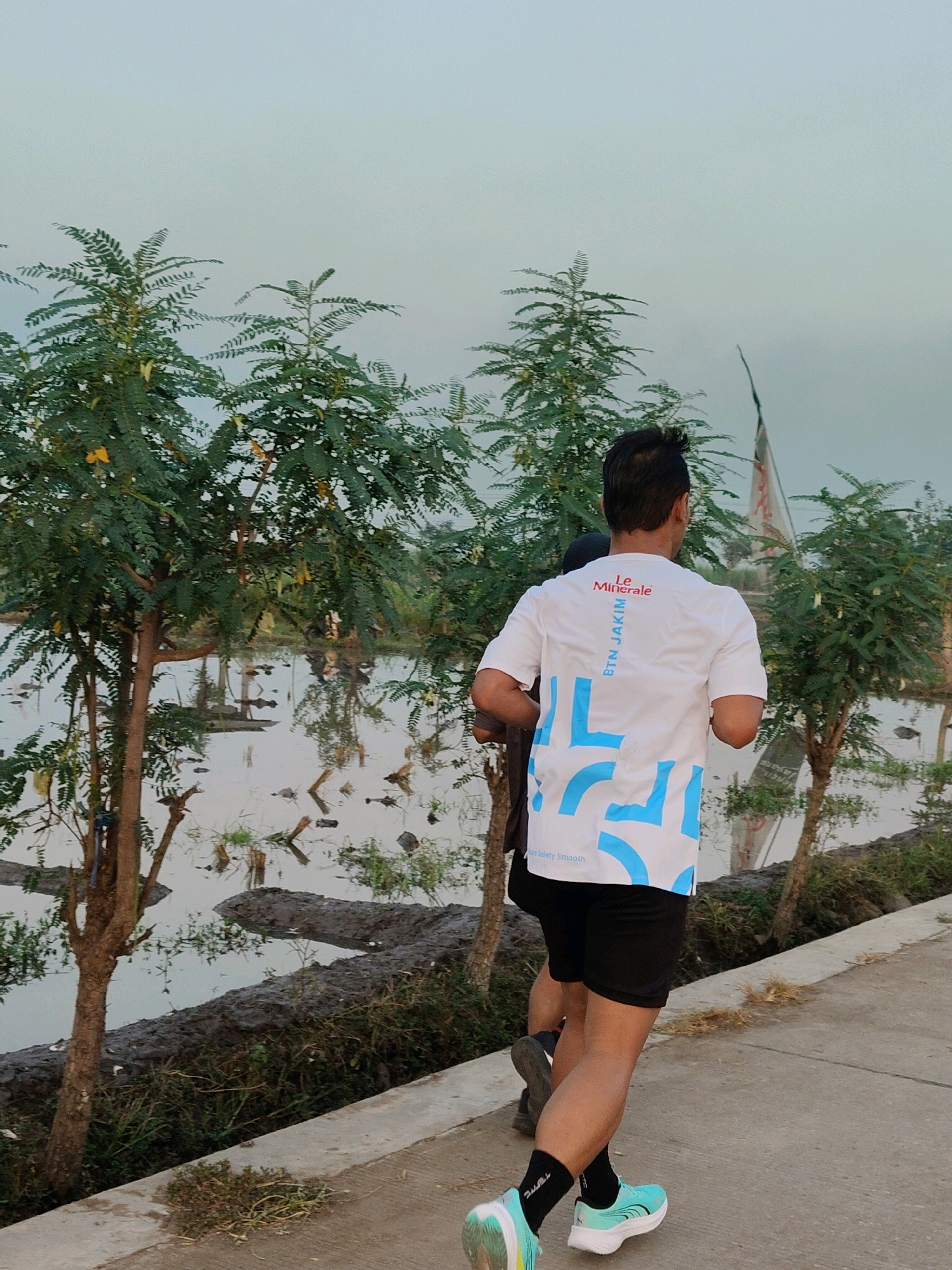 In Sumengko, Indonesia, two individuals are captured from behind as they run on a concrete path beside a body of water. The person in the foreground, wearing black shorts and a white t-shirt with blue geometric patterns and text that reads "Le Minerale" and "BTN JAKIM," is in motion. Their black socks and teal running shoes are visible. A second runner, partially obscured, is running slightly ahead and to the left. The environment includes several trees with green foliage and yellow flowers, and the background shows a tranquil body of water reflecting the sky and trees. The sky is overcast with a pale blue-grey hue. The water is calm, with debris and vegetation visible on its surface and along the muddy banks.