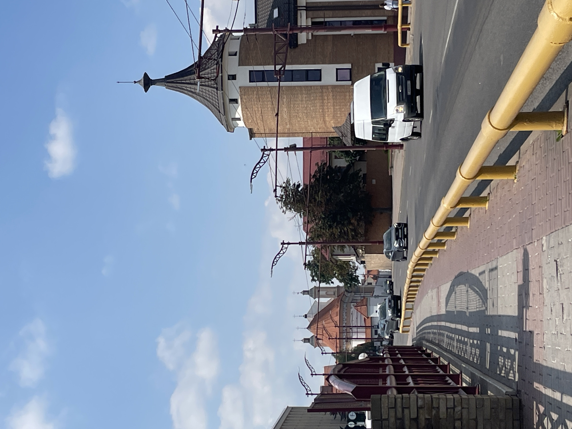 This is a street-level photograph taken on a sunny day in Hrodna, Belarus, depicting a road with a pedestrian walkway and buildings. The perspective is from a slightly elevated position, looking down a paved road.
On the right side of the road, there is a distinctive multi-story building with a conical, dark-tiled roof topped with a spire. The building features light brown brickwork and sections with white facade, and it has several tall, narrow windows. A white van is driving on the road in front of this building. Further down the road, other vehicles, including dark-colored cars, are visible.
On the left side of the road, a red metal fence runs along the edge of a pedestrian walkway. This walkway is paved with interlocking bricks in shades of red and gray. The walkway is bordered by a bright yellow metal railing. Behind the fence, a light-colored building with a stone facade is partially visible. In the background, several church buildings with orange-tiled roofs and towers can be seen. Trees are also present in the scene. The sky is blue with scattered white clouds. A person is visible walking on the sidewalk in the far right of the image.