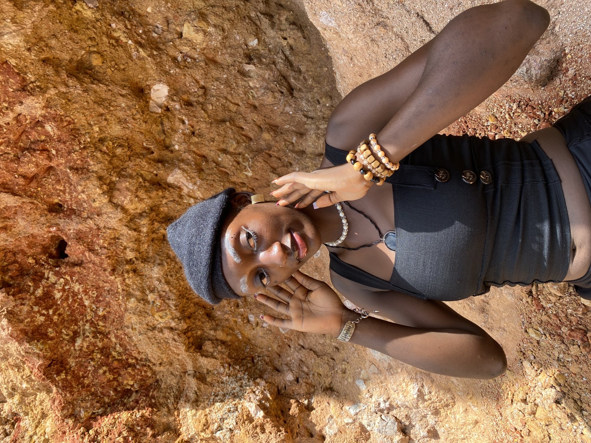 A young Black woman is posing in front of a rocky, reddish-brown cliff face. She is wearing a black beret, a black tank top, and dark pants. She has white makeup on her eyebrows and a white line of eyeliner above her eyes. She is also wearing several beaded bracelets and necklaces, and large gold hoop earrings. Her hands are raised to her face, with her fingers spread. The lighting suggests it is daytime, with shadows cast by the sun. The location is identified as Bar Jirgi Summa, Nigeria.