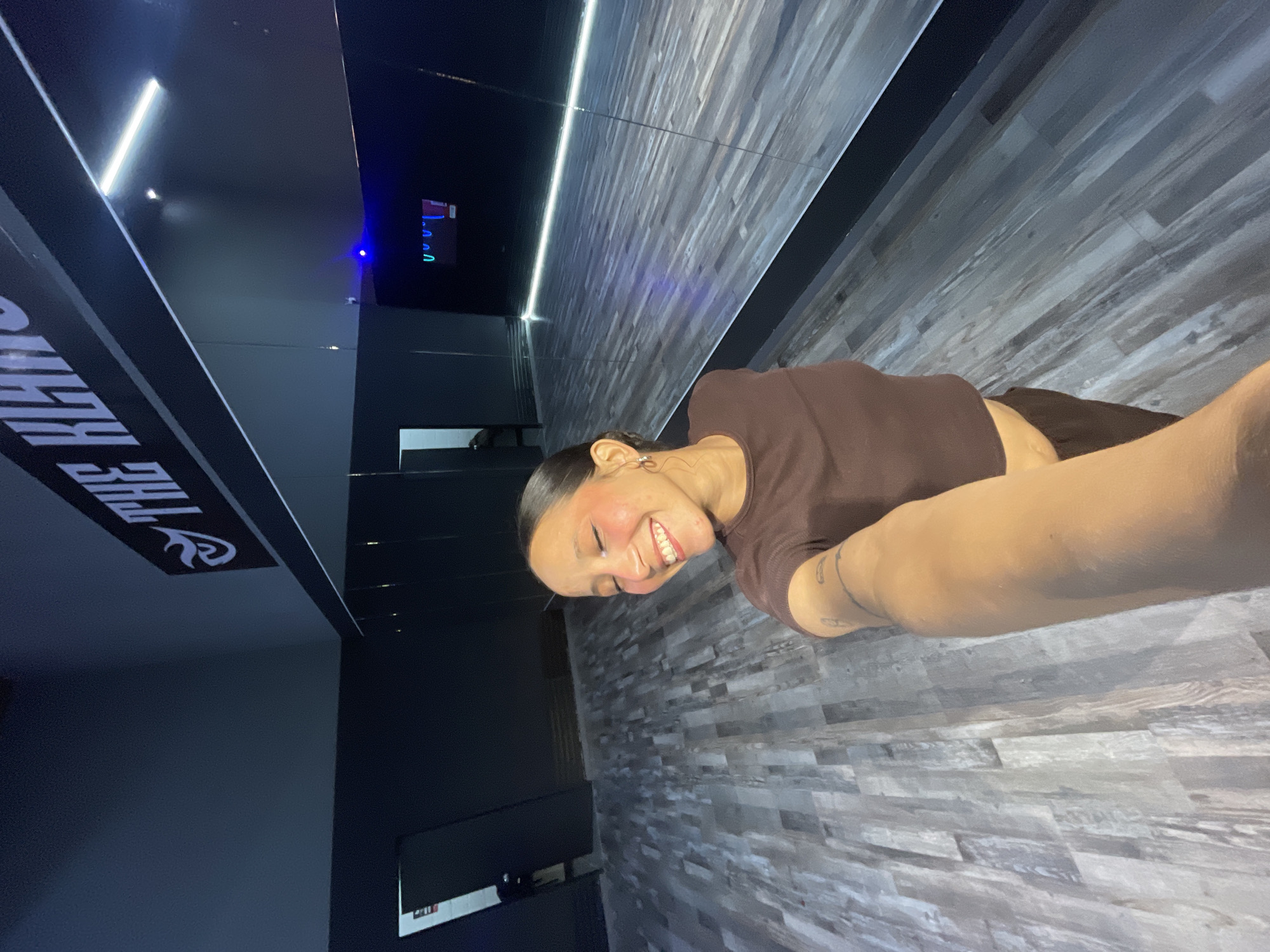 A woman is taking a selfie in what appears to be a dance studio. She is smiling and has her eyes closed. She is wearing a brown crop top and dark pants. The studio has a wooden floor and mirrors on one wall. There is a sign on the wall that says "THE KHAN". The lighting is dim, with some neon lights in the background. The woman has dark hair pulled back in a ponytail and is wearing hoop earrings. She has blush on her cheeks. The studio is located in Pampatar, Venezuela.