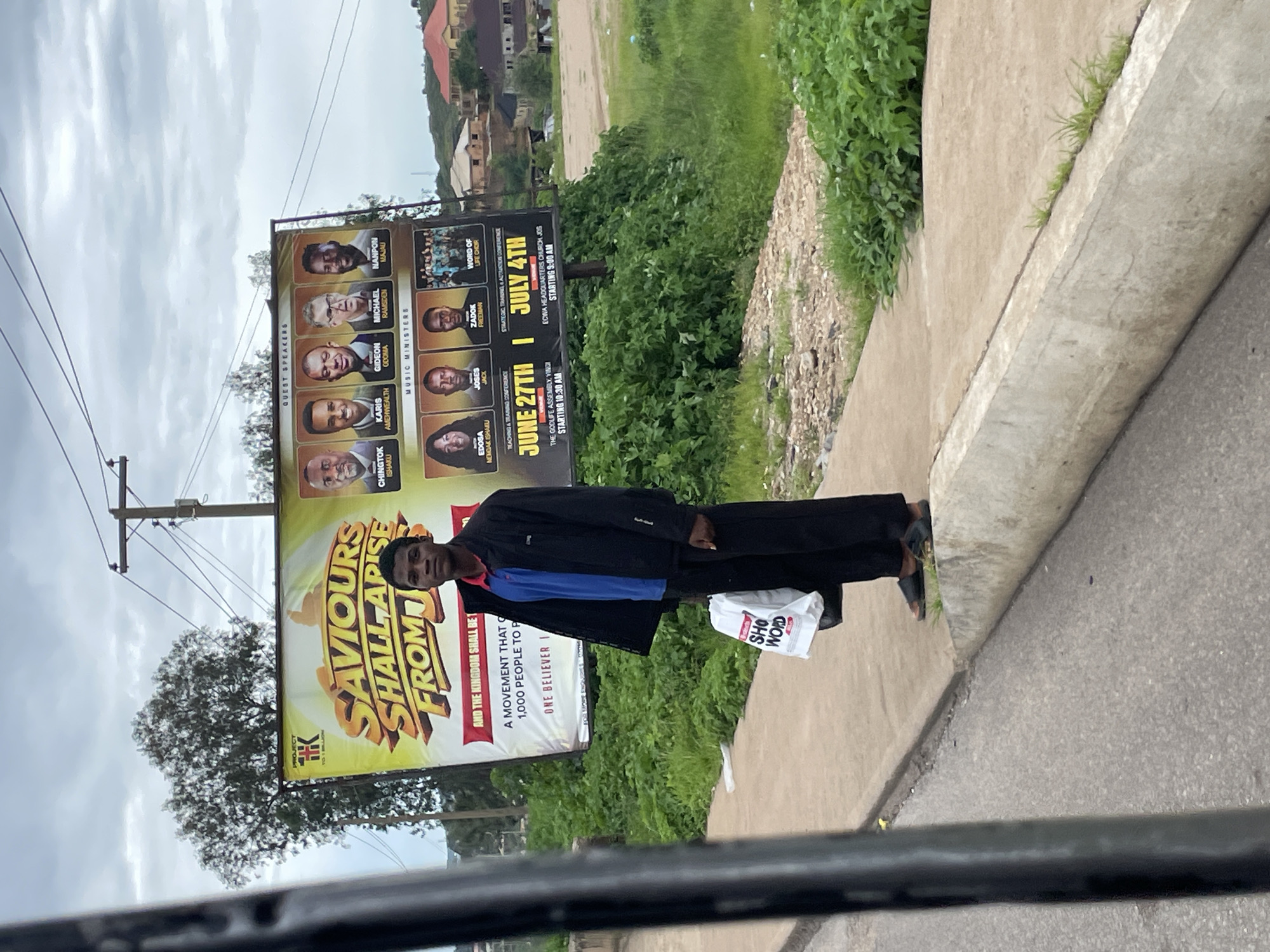 A young Black male stands on a sidewalk in Gura Top, Nigeria, beside a large billboard. The billboard advertises an event called "Saviours Shall Arise From Us" featuring "Quest Speakers" and "Music Ministers." The man is wearing a dark jacket over a blue and red collared shirt and dark pants. He is holding a white shopping bag with the words "SHo WORD" printed on it. The background shows lush green foliage, trees, and some residential buildings in the distance, under an overcast sky. A utility pole with wires is visible behind the billboard. The sidewalk is made of concrete, and a drainage ditch runs along its edge.
