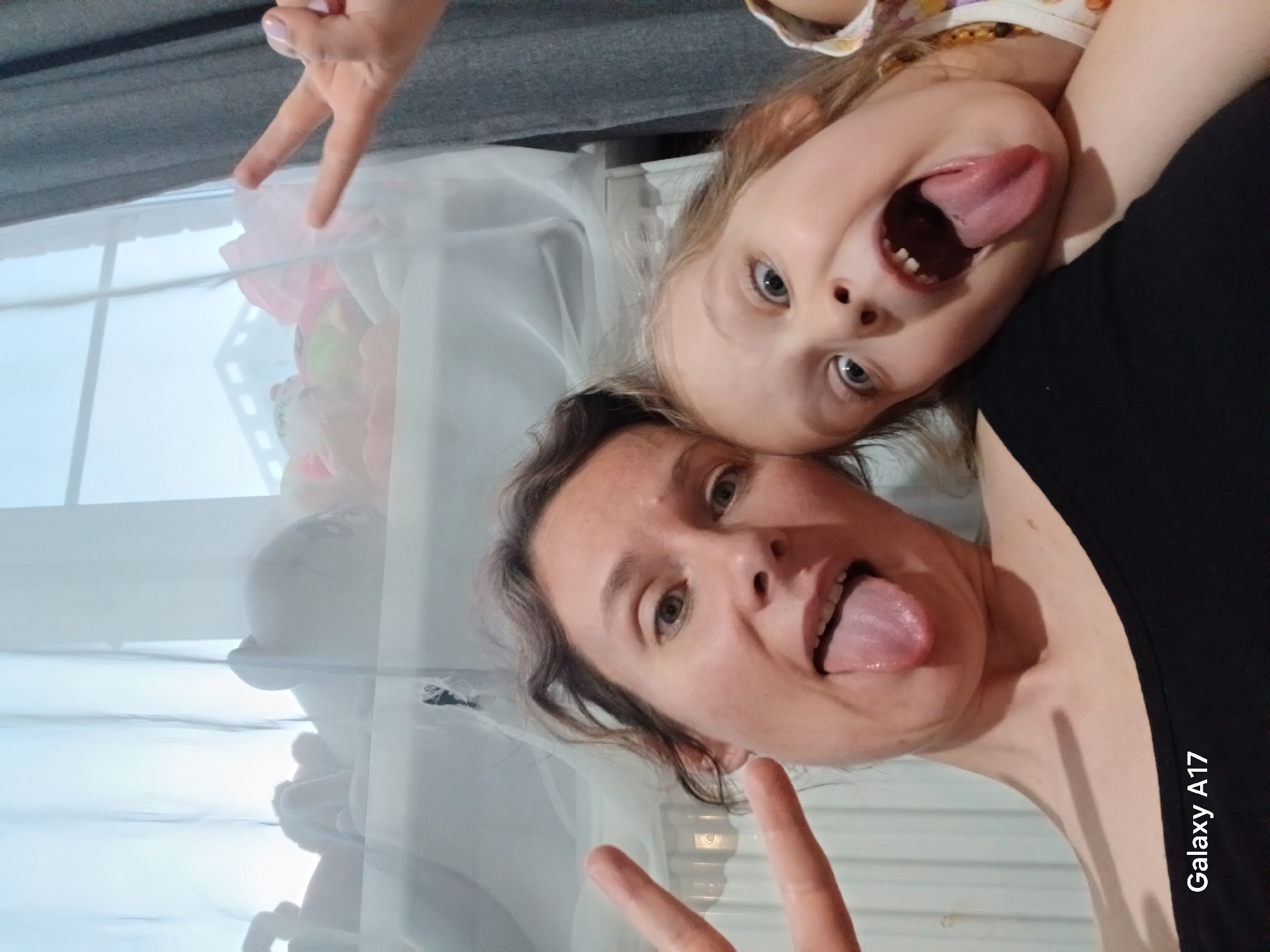 A selfie, taken indoors, depicts a light-skinned woman and a young light-skinned child with blond hair. Both are sticking out their tongues. The woman is wearing a black top. A peace sign is formed with fingers near the bottom of the frame. In the background is a white curtain, possibly a window, and some stuffed toys. The phone "Galaxy A17" watermark is visible on the right side. The location is inferred to be Yekaterinburg, Russia, only from the prompt and no elements within the image support this.