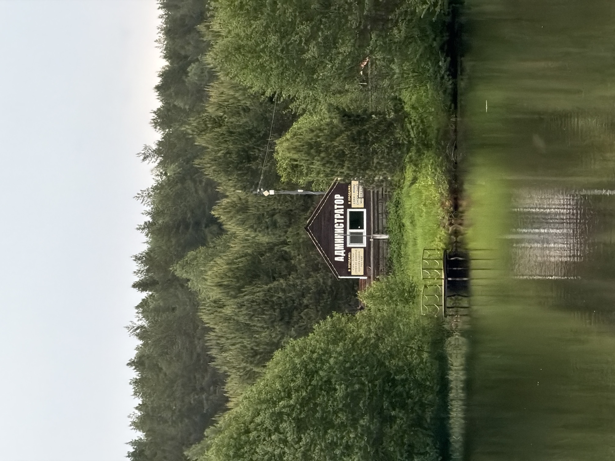 A landscape photograph depicts a body of water with the edge of a forest to the left, and an administrator's building nestled among the trees, possibly located in Prikamskii, Russia. The administrator's building has a sign in Cyrillic, with the text АДМИНИСТРАТОР vertically aligned, along with an advertisement that mentions products for sale such as sunflower seeds, bait, and fish-related items. The edge of the water has a small dock or pier that extends into the water with some grassy area adjacent to the building. A tall pole with what appears to be some sort of electrical or communication equipment is located near the building. The overall lighting suggests an overcast day. The water surface reflects the sky and the trees.