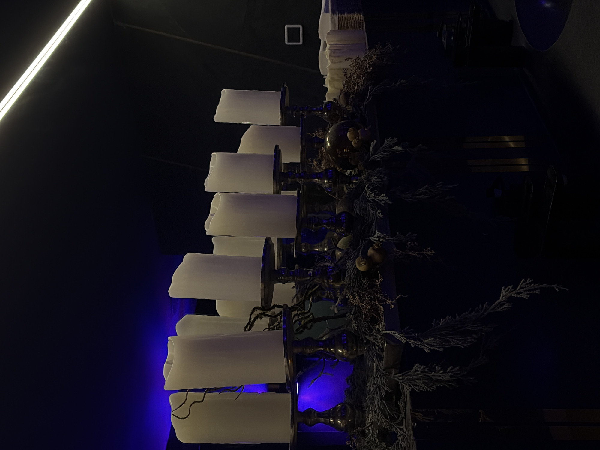 The image displays an interior scene, likely a commercial establishment such as an upscale lounge, bar, or restaurant, located in Moscow, Russia. The precise address or venue name is not identifiable from the visual information provided.
The primary subject is a decorative arrangement situated on a dark, flat surface, resembling a counter or bar. This arrangement features approximately 10-12 tall, white, pillar-style artificial candles, each set atop an ornate metallic candle holder, appearing to be brass or bronze-toned, with varying heights and sculpted bases. The candles exhibit an uneven, melted-wax-like top but lack visible flames, consistent with LED or battery-operated units. The base of this display is adorned with a dense layer of frosted or dried evergreen foliage, interspersed with dark, gnarled twigs and small, spherical, light-brown decorative elements resembling miniature gourds or ornamental fruits.
The background is dimly lit, with a prominent section to the left bathed in a deep blue-purple ambient light. A bright, linear white light fixture, possibly an LED strip or fluorescent tube, is visible diagonally across the upper right ceiling. To the right, a small, white, rectangular object is mounted on a dark wall, and a woven basket containing folded white items (likely napkins or towels) rests on an adjacent surface. The environment is dark and modern, creating an atmospheric, sophisticated ambiance. No individuals, specific actions, or interactions are present in the scene.