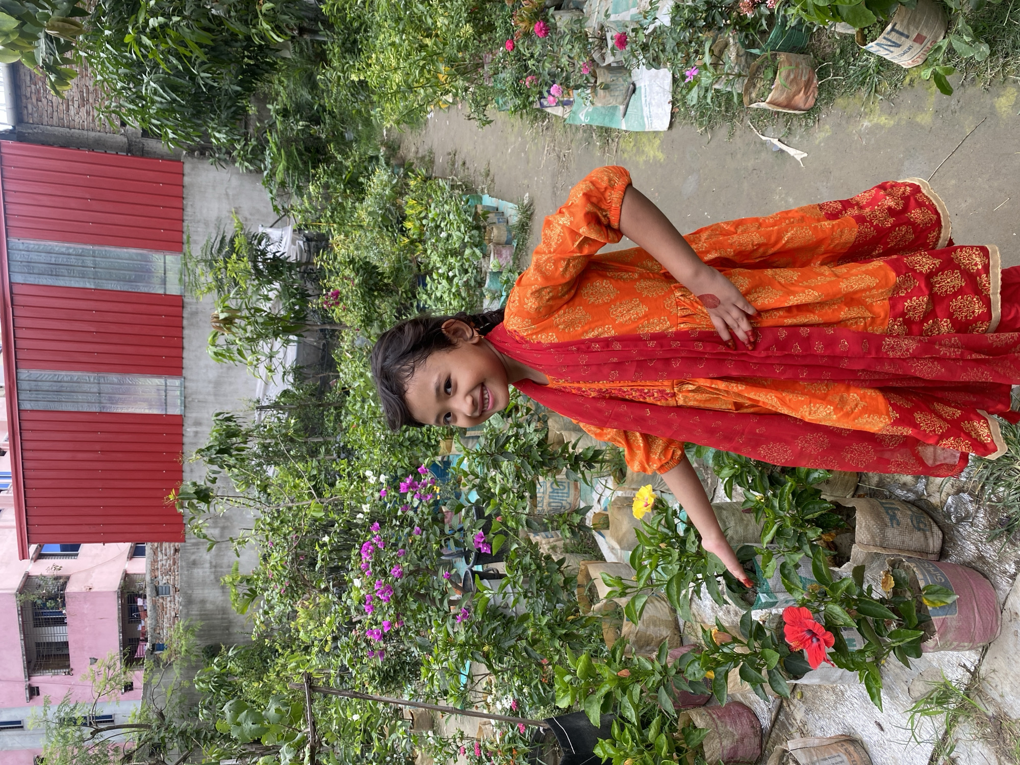 A young girl in an orange and red dress with gold patterns is outside tending to flowers. She is slightly bent over with her hand reaching toward a red hibiscus flower. She is smiling at the camera. The flowers are in bags marked with "Cement". The background includes other flowers, plants, and parts of buildings. The city is Dhaka, Bangladesh.