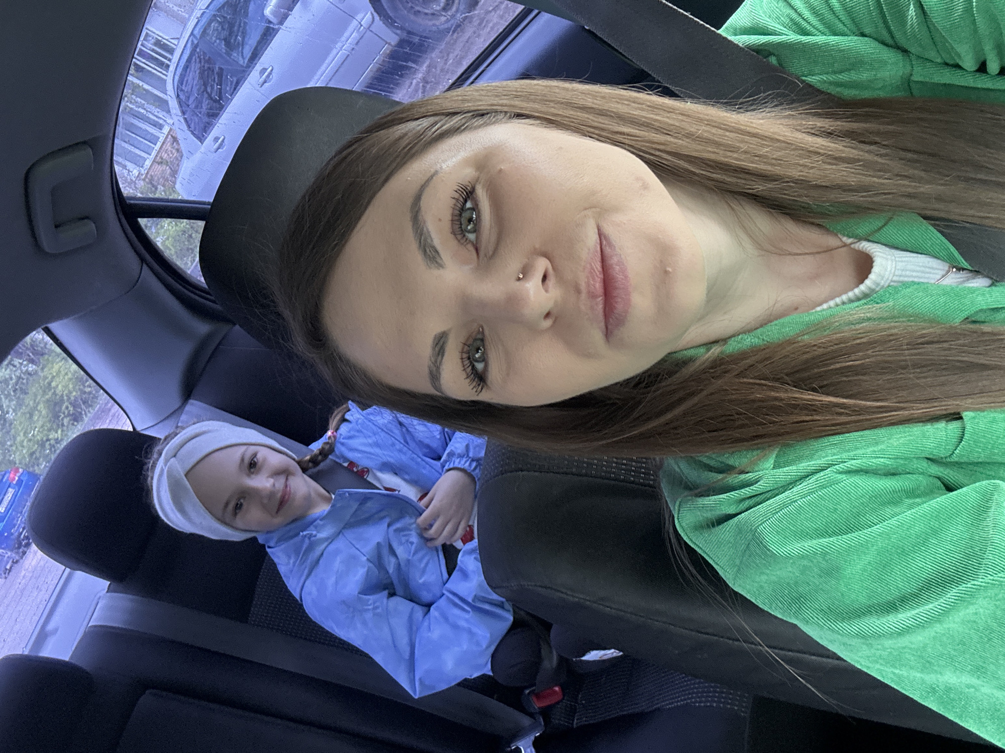 A selfie taken inside a car shows a young woman in the front seat wearing a green jacket and seatbelt. A young girl in the back seat wears a blue jacket and a white headband. The background shows the outside environment through the car window. While the location is identified as Razdolye, Russia, specific details within the scene are not clearly discernible to confirm this.