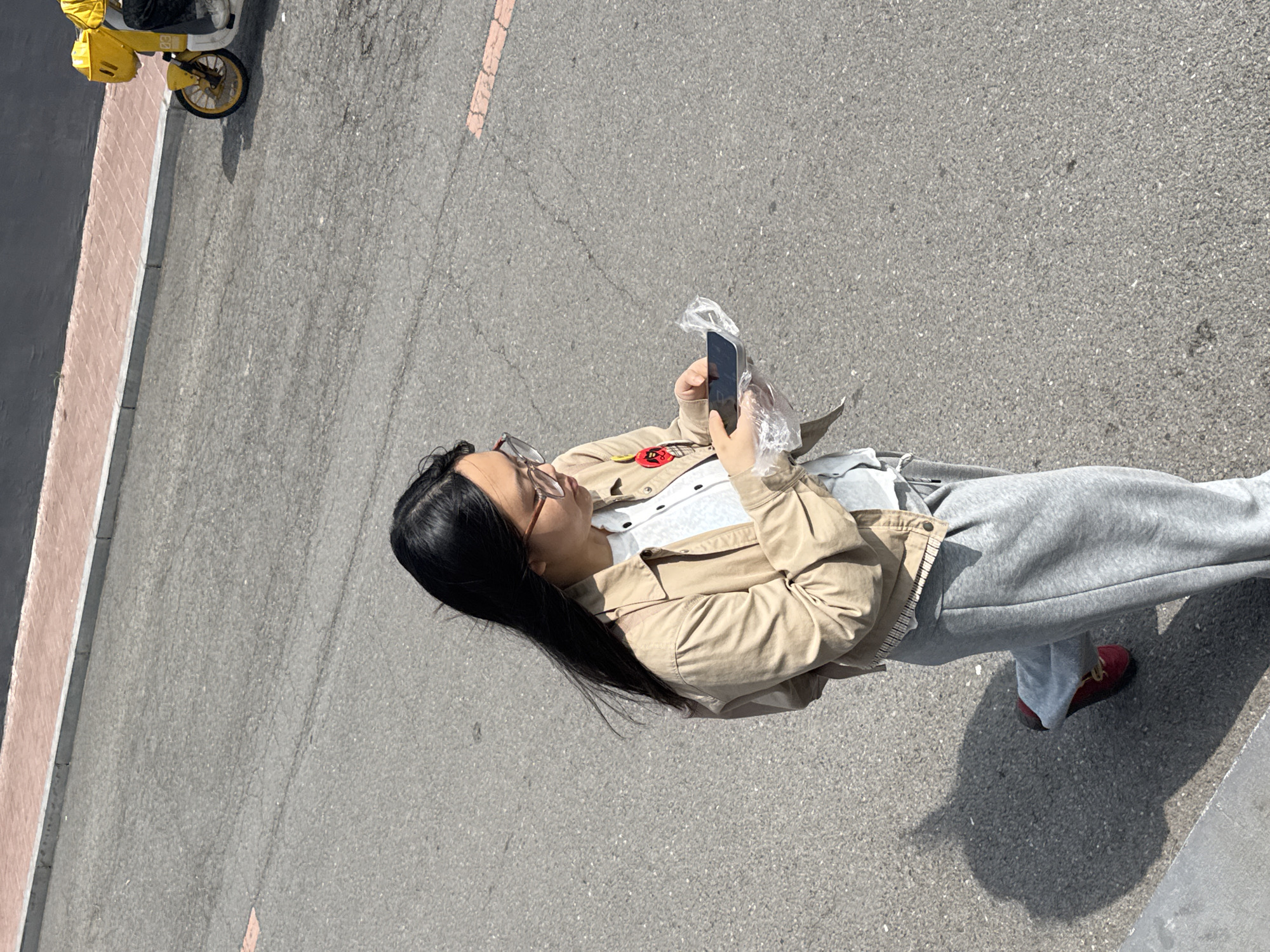 A high-angle, full-body shot captures a young person, likely female, standing outdoors on a paved surface in Zhongtun, China. She has long, straight black hair and wears glasses. She is dressed in a light tan or beige long-sleeved jacket over a white top, grey sweatpants, and red shoes with yellow laces. A circular red pin with an indistinct white symbol is affixed to her jacket.
The individual is actively engaged with a smartphone, holding it in her right hand. The phone appears to be partially covered or wrapped in a clear plastic bag. Her head is tilted downwards, indicating focus on the device.
The scene is an outdoor environment, likely a street, path, or parking area, characterized by a grey asphalt surface. To the left, there is a curb separating the main paved area from a darker section of pavement. In the upper left background, a portion of a yellow scooter or bicycle is visible, showing a wheel and a segment of its body, which features a faint "03" logo.
The photograph is taken during bright daylight hours, evidenced by strong, distinct shadows cast on the ground by the person and other objects, suggesting a sunny and clear day. The subject's expression is focused on her phone, without discernible strong emotions.