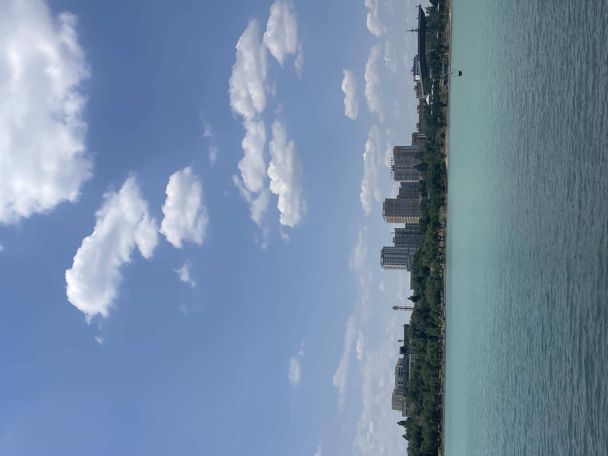 The image depicts a wide, calm body of water with a turquoise hue in the foreground. In the middle ground, a lush green tree line runs along the horizon, separating the water from a cityscape. The cityscape features several modern, multi-story buildings, some appearing to be residential or office complexes, with a few industrial structures and a tall red and white pylon visible. The background is dominated by a clear blue sky with scattered fluffy white clouds. In the water, a small black buoy with a white top is visible. The scene appears to be a waterfront area in a city, with a blend of natural landscape and urban development. Based on the architectural style of the buildings and the presence of the large body of water, this scene is likely located in Almaty, Kazakhstan, possibly along the Small Almaty Gorge Reservoir, also known as the Almaty Arasan.