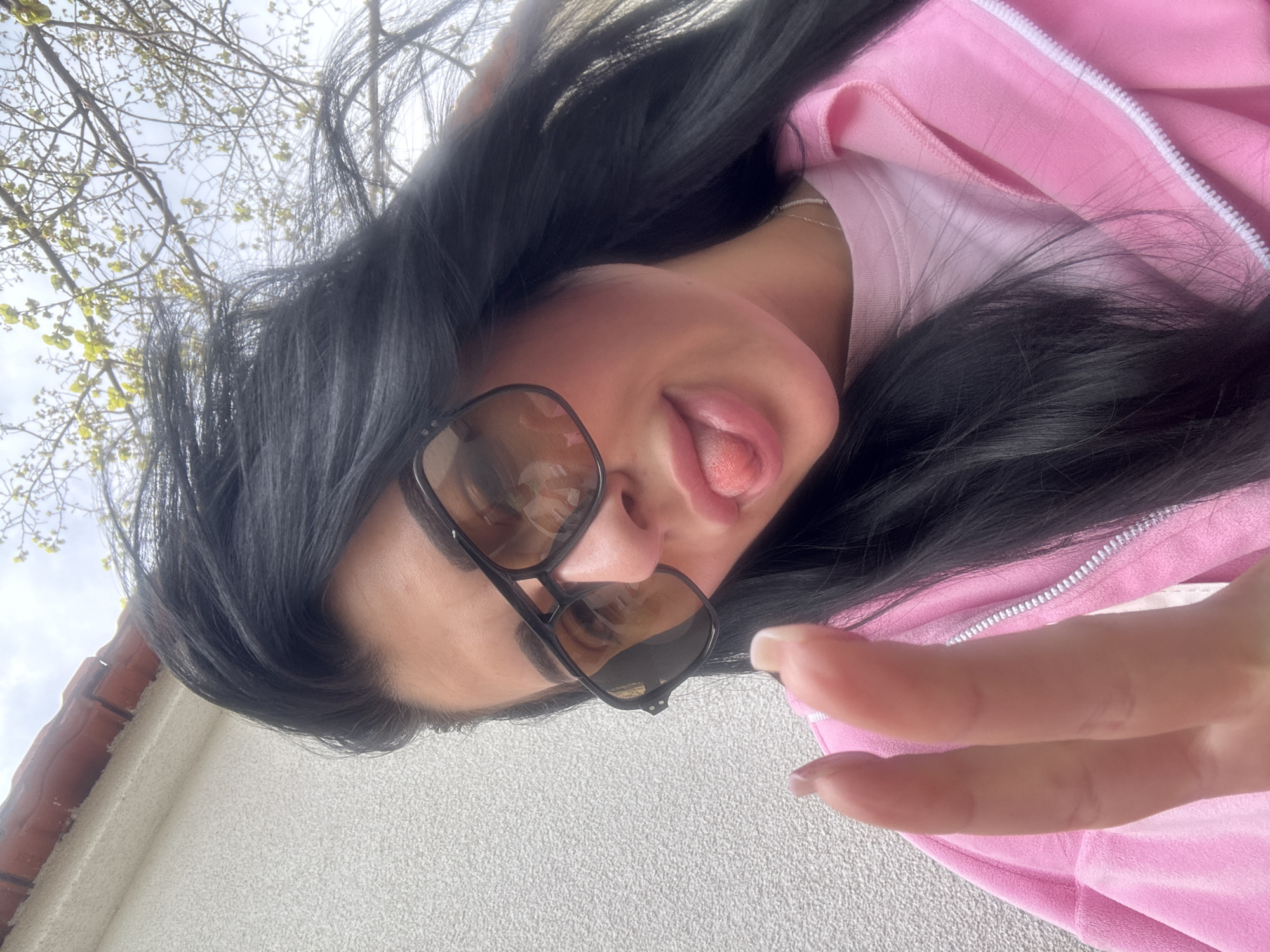 A person with dark hair, wearing sunglasses and a pink jacket, is posing for a selfie while sticking their tongue out. The background includes a building with a red brick trim and tree branches. Based on the location provided, it is likely in Saedinenie, Bulgaria.