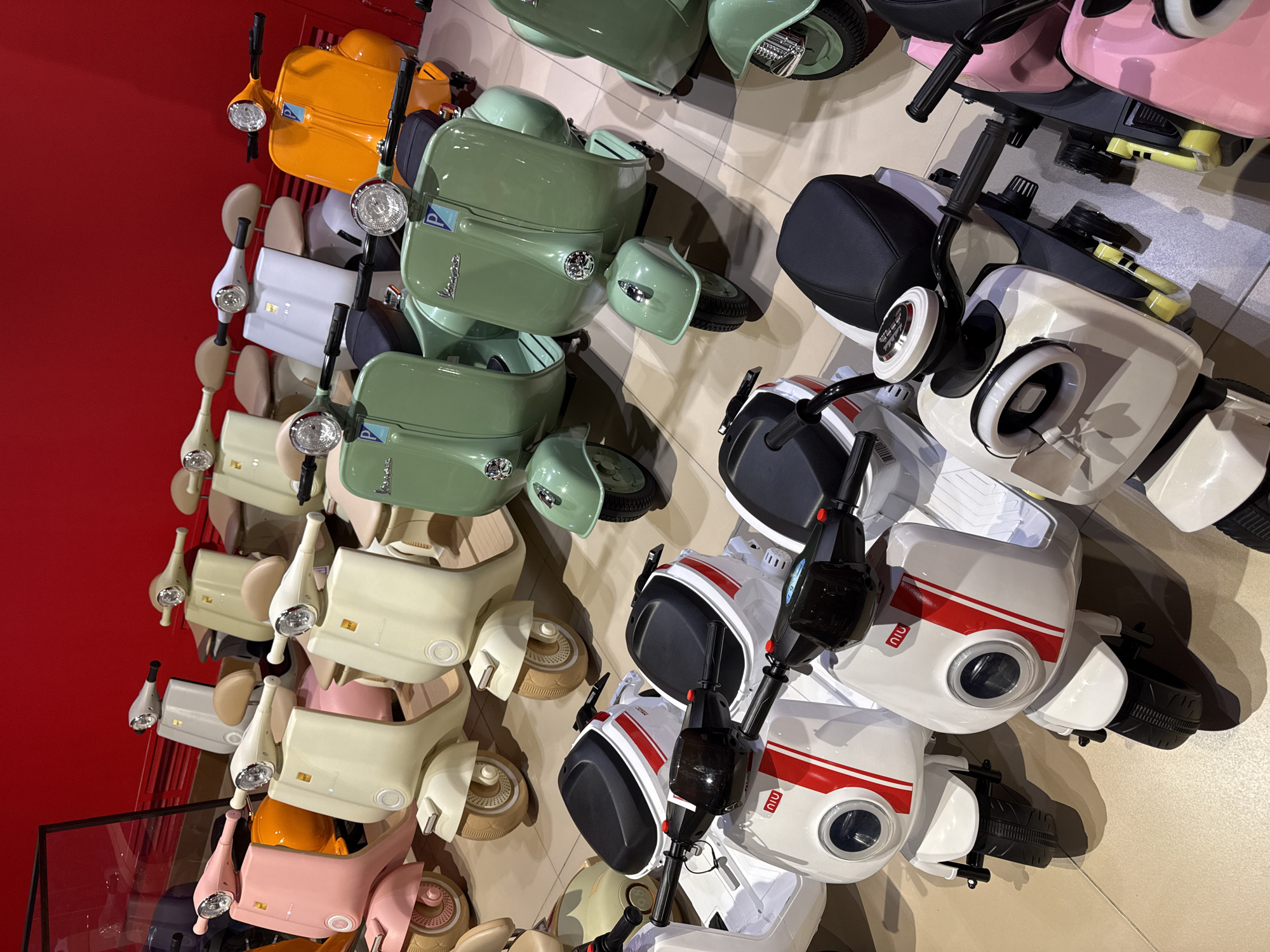 The image shows a display of children's toy scooters, arranged on a wall, likely for sale in a retail setting. The scooters are of varying colors, including orange, green, white, beige, and pink. Some models appear to mimic classic Vespa designs, while others are more modern, such as the white models bearing the "NIU" logo. The scooters are positioned closely together, creating a dense collection of items. The background includes a red wall on the left and a tiled floor on the right. This image was taken in Moscow, Russia.