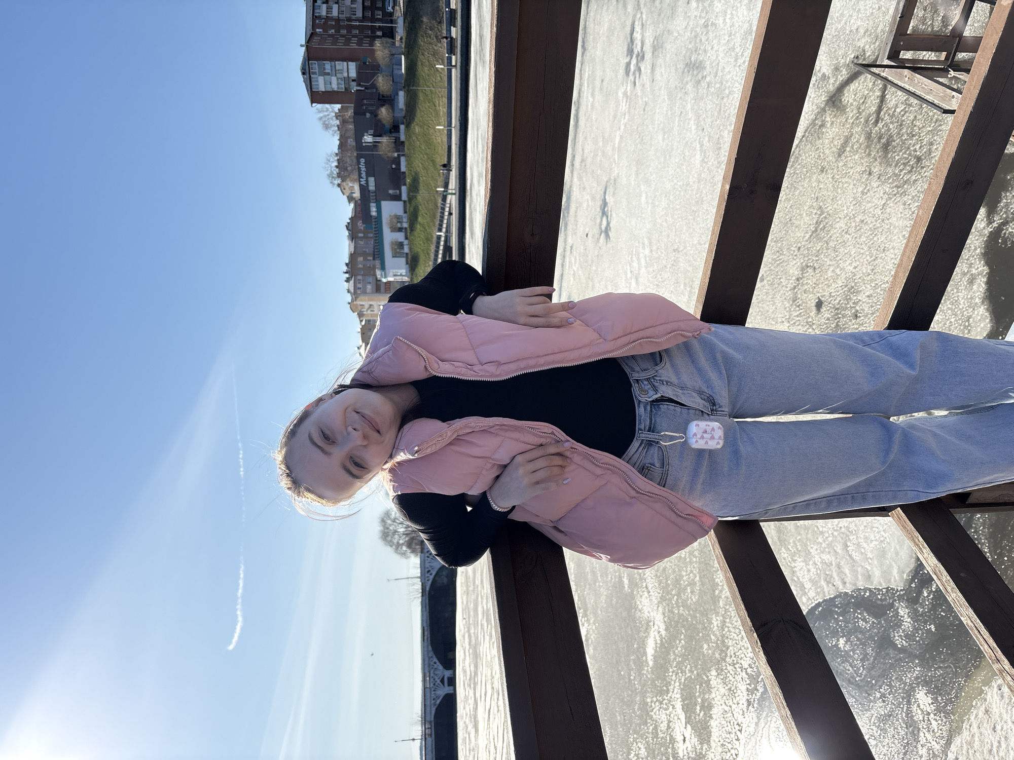 A young woman is captured in a bright outdoor portrait, leaning on a dark wooden railing. She has light-colored hair and a gentle smile, looking directly at the camera. She is wearing a light pink puffer vest over a black long-sleeved top and light-wash blue jeans. A white AirPods case with a subtle pattern is clipped to the belt loop of her jeans.
The setting appears to be a boardwalk, pier, or bridge overlooking a body of water, possibly a river or lake, that is frozen or covered in ice, glistening under the sun. In the background, an urban landscape features several multi-story buildings and a distinct, multi-arched bridge. A sign on one of the distant buildings partially reads "Maestro."
The weather is sunny and clear, indicated by the vibrant blue sky, strong sunlight causing lens flare, and a faint contrail from an aircraft. The time of day is daytime. The overall mood is calm and relaxed, suggesting a casual outing on a bright, possibly chilly day, consistent with the location context of Votkinsk, Russia.