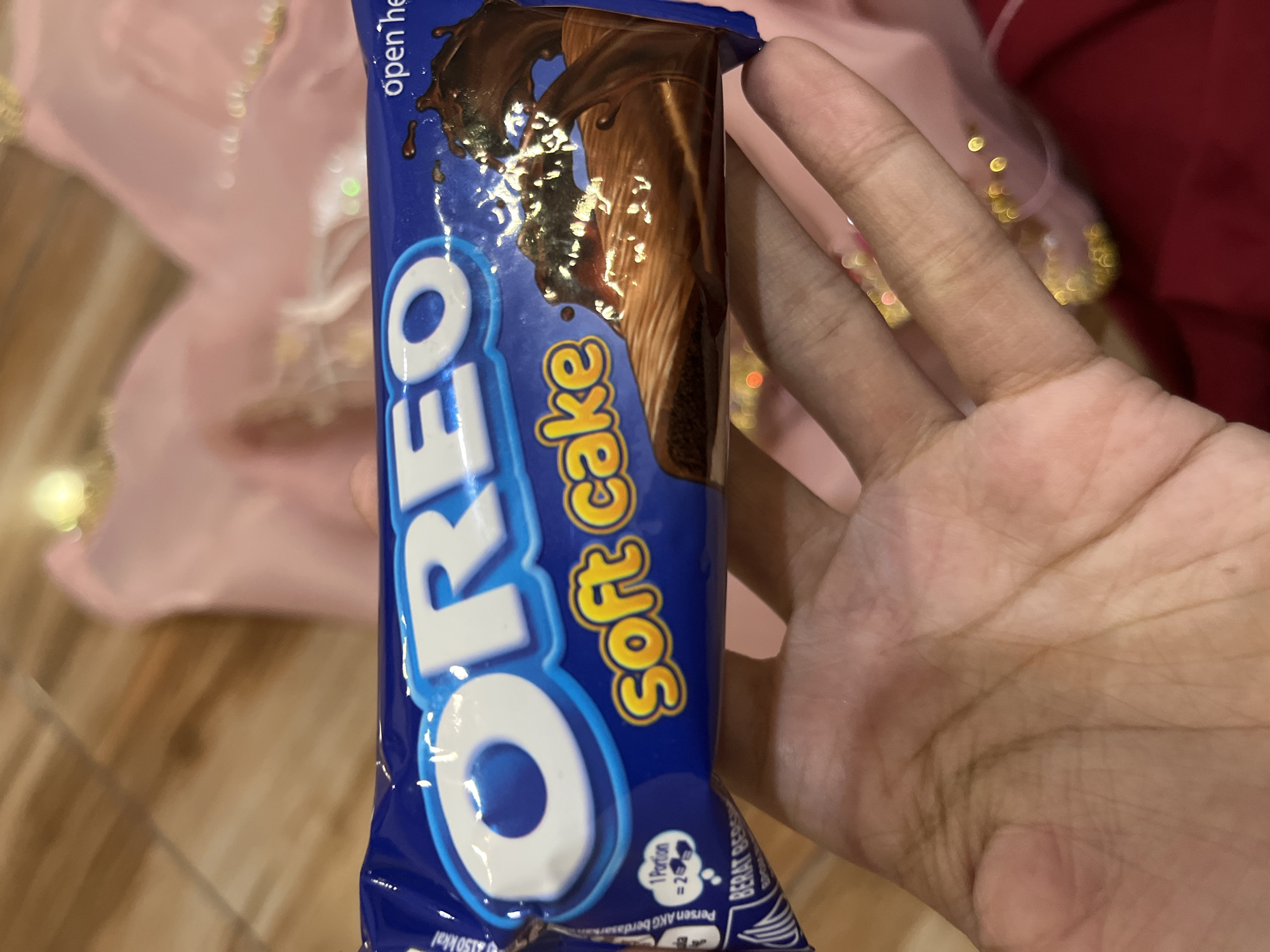 An overhead, close-up shot captures an Oreo soft cake snack package being held in a person's hand. The package is a vibrant blue with white and yellow text. The Oreo logo is prominent, and below it reads "soft cake" in yellow lettering. A chocolate cake with chocolate frosting is depicted on the right side of the package. The hand holding the package appears to be of a lighter skin tone with visible palm lines. The background is blurred and shows a light pink fabric and some out-of-focus golden or yellow lights. The scene is indoors, with the flooring appearing to be a wooden tile. The image is not indicative of any specific location in the city of Palopo, Indonesia, or any other city.