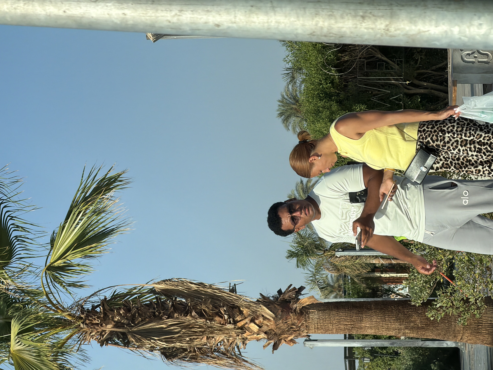 In Sharm El Sheikh, Egypt, a man and a woman are standing outdoors near a palm tree under a clear blue sky. The man, wearing sunglasses, a light grey t-shirt, and grey sweatpants with "alo" branding, holds a mobile phone in his left hand and appears to be looking at it. The woman, with her hair in a bun, wears a yellow sleeveless top and a leopard-print skirt. She is holding a mobile phone in her right hand, and a black clutch bag is attached to her left hip. Both are looking down at their phones. In the background, lush green foliage and other palm trees are visible. A metal pole is present in the foreground on the right side of the frame.