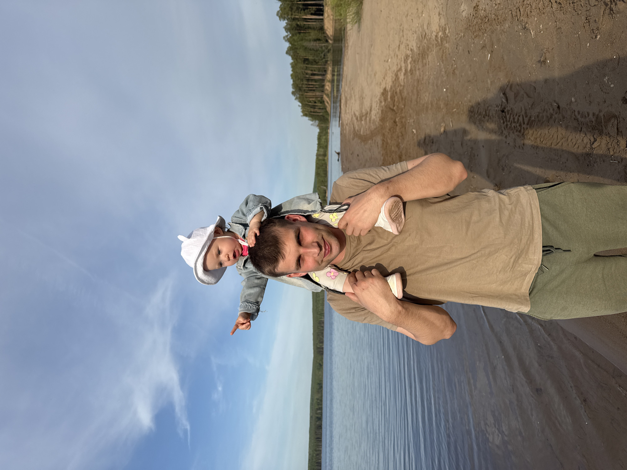 On the shore of a body of water in Chaikovsky, Russia, a man is standing with a baby on his shoulders. The man is wearing a beige t-shirt and green pants. The baby is wearing a light blue jean jacket and white hat. The baby is pointing towards the sky. Behind the man is the water's edge, with the water stretching into the distance. A beach borders the water's edge, which then turns into trees. The sky is blue with some scattered clouds.