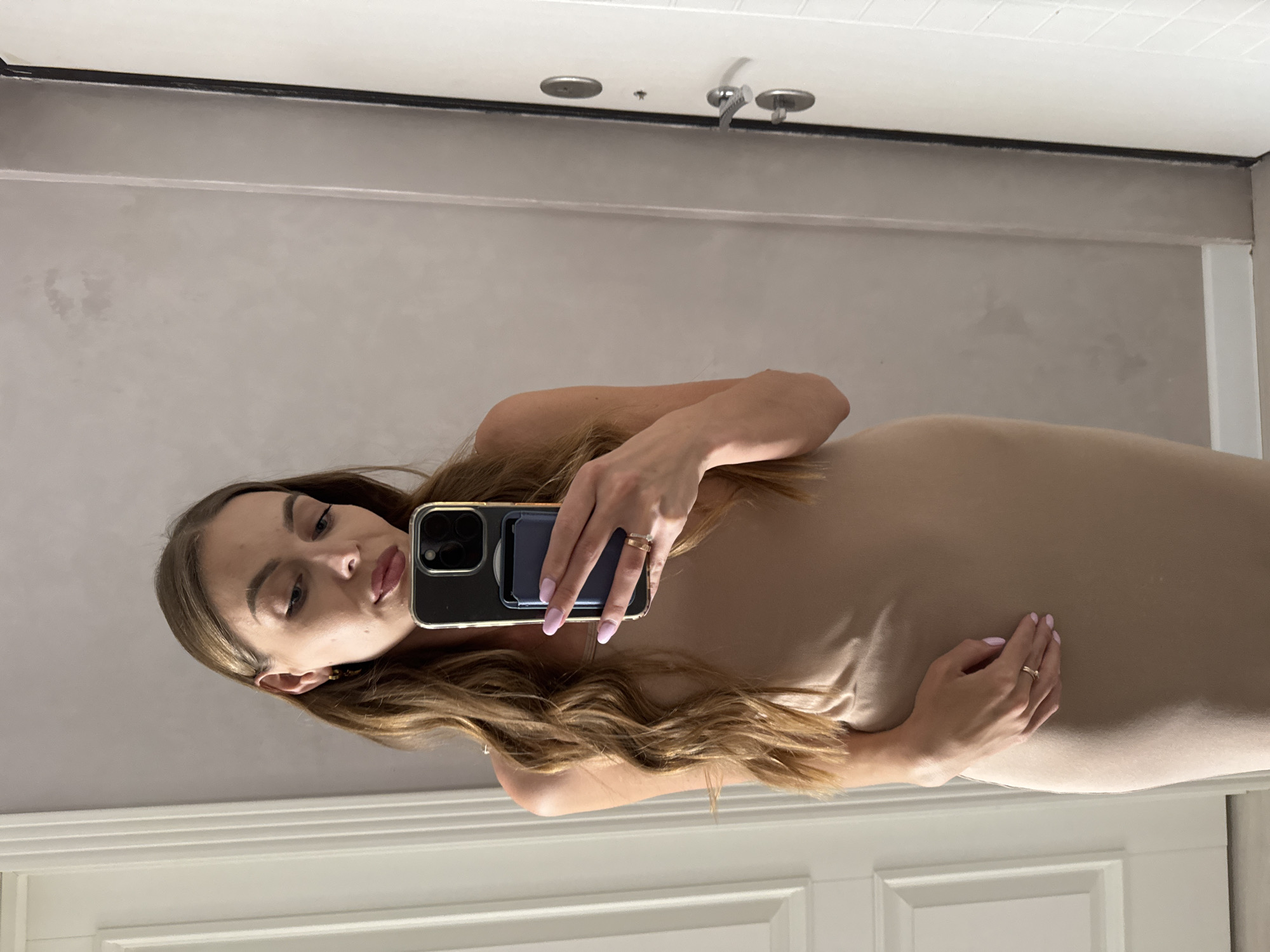 A young woman with long, wavy brown hair is taking a selfie in a mirror. She is wearing a form-fitting, sleeveless beige dress. She is holding a smartphone with a gold-colored case and a blue card holder attached to the back. Her left hand is resting on her stomach, and she is wearing a ring on her left ring finger. Her nails are painted a light pink color. The background appears to be an indoor setting, possibly a home, with a white door on the left and a white door with a silver handle on the right. The wall has a textured, light gray finish with some subtle discoloration. The image does not contain any specific visual cues to indicate the location is Sochi, Russia.