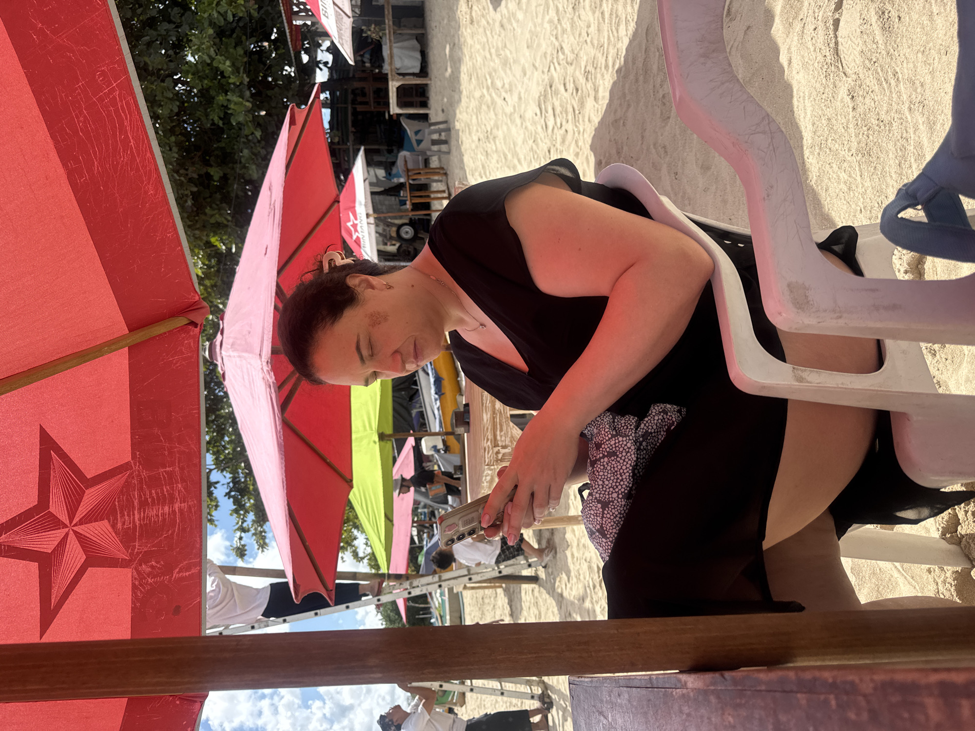 A woman with dark hair tied back sits on a white plastic chair on a sandy beach, looking down at her mobile phone. She wears a black top with short sleeves. The chair is positioned under a large red umbrella with a star design. In the background, other beach umbrellas of red and green are visible, along with some tables and chairs. Several people are in the background, including someone on a ladder. The location is Kuta Selatan, Indonesia.