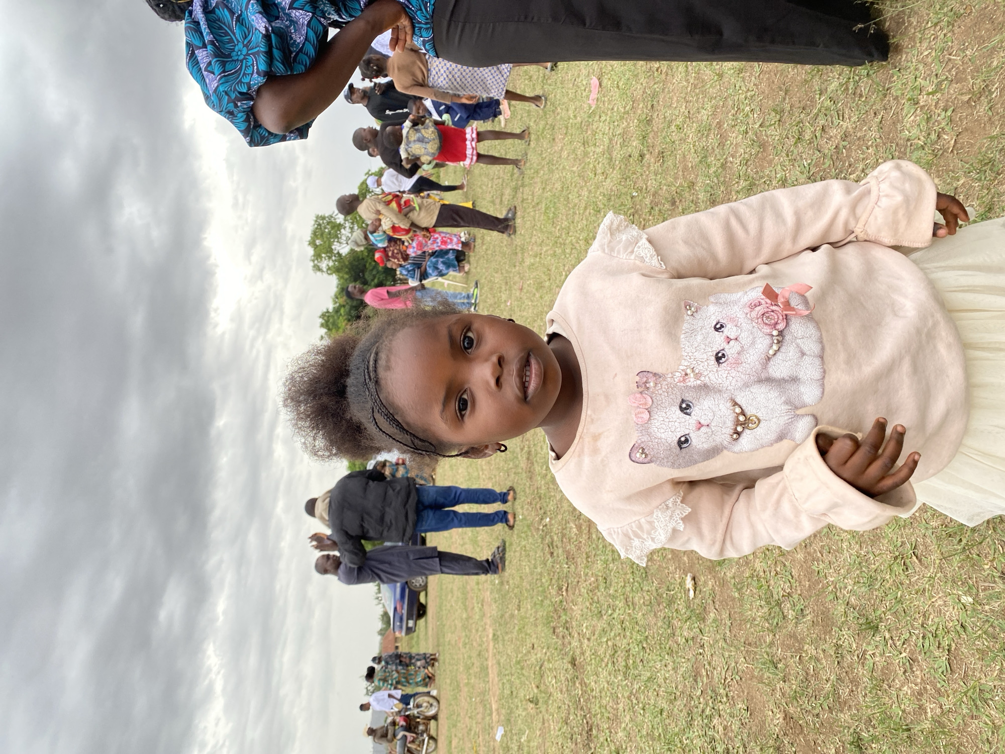 The image depicts a young African girl in Kuru Dungus, Nigeria, standing on a grassy field amidst a crowd of people. The girl is wearing a light-colored long-sleeved shirt with a cat graphic and a light-colored skirt. The crowd of people in the background consists of adults and children. Some are dressed in casual attire, while others are in more formal clothing. The background also shows a couple of cars and motorcycles. The sky is overcast.