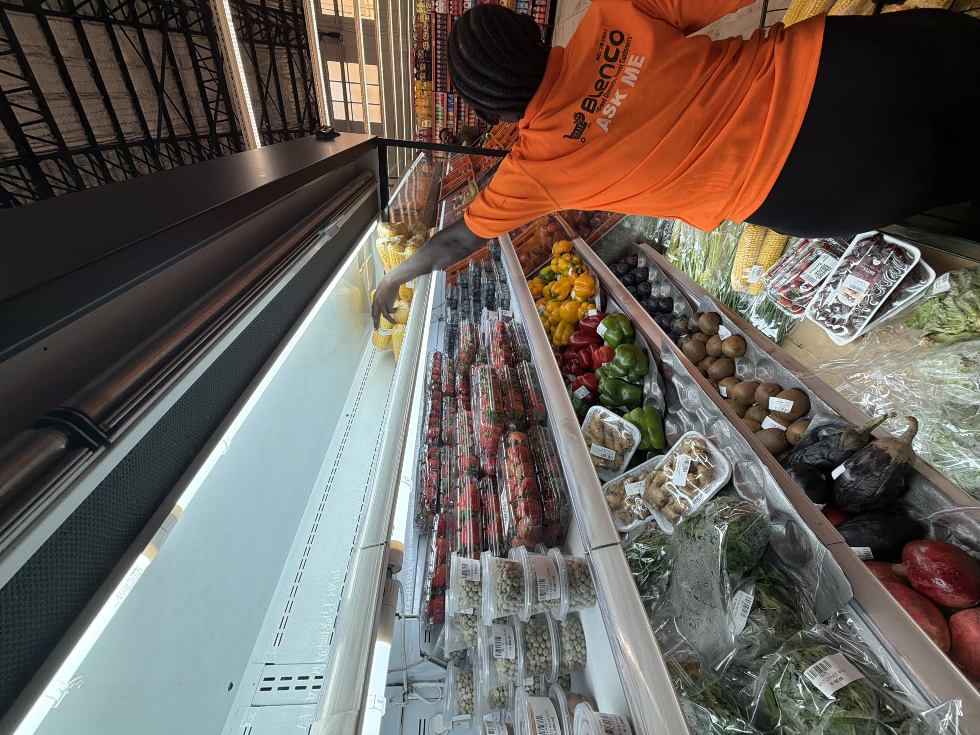 The image depicts a person in a bright orange shirt with "Blenco" printed on it, reaching into a refrigerated display case stocked with various fruits and vegetables in a store. The person is likely a worker at Blenco supermarket. The display case contains items like corn on the cob, blueberries, strawberries, yellow bell peppers, red bell peppers, green bell peppers, kiwis, eggplants, and other greens. Based on the OCR text, the setting is likely the city of Igboefon, Nigeria, where Blenco supermarkets are located.