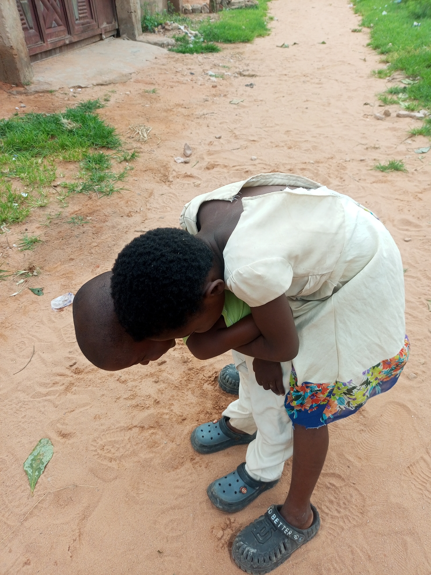 Two young children are engaged in an interaction on a sandy path in Tempure, Nigeria. The child in front, with dark curly hair, is bent over, their head down. They are wearing a light green shirt and light-colored pants, and dark blue clogs. The second child, mostly obscured, is standing and has their arms around the first child. This child is wearing a cream-colored dress with a floral hem and white pants, also wearing dark blue clogs. The environment consists of a sandy ground with patches of dry grass and some scattered debris. In the background, a section of a building with a dark wooden door is visible, and a path recedes into the distance, bordered by grass.