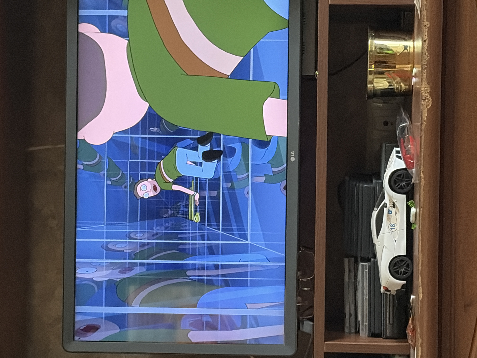 In an indoor setting, a television displays an animated scene from Rick and Morty. The scene shows multiple versions of Morty Smith within a grid-like environment. One Morty is in the foreground, seen from behind, wearing a green t-shirt with brown and white stripes and blue pants. Another Morty is in the middle ground, facing the camera with a surprised expression, holding a skateboard. The background is a blue grid with reflections of more Mortys. Below the television, on a wooden shelf, sits a white toy sports car, and to the right, a gold-colored cylindrical container. Various DVDs are stacked on the left side of the shelf. A pair of glasses rests on the top edge of the television stand.