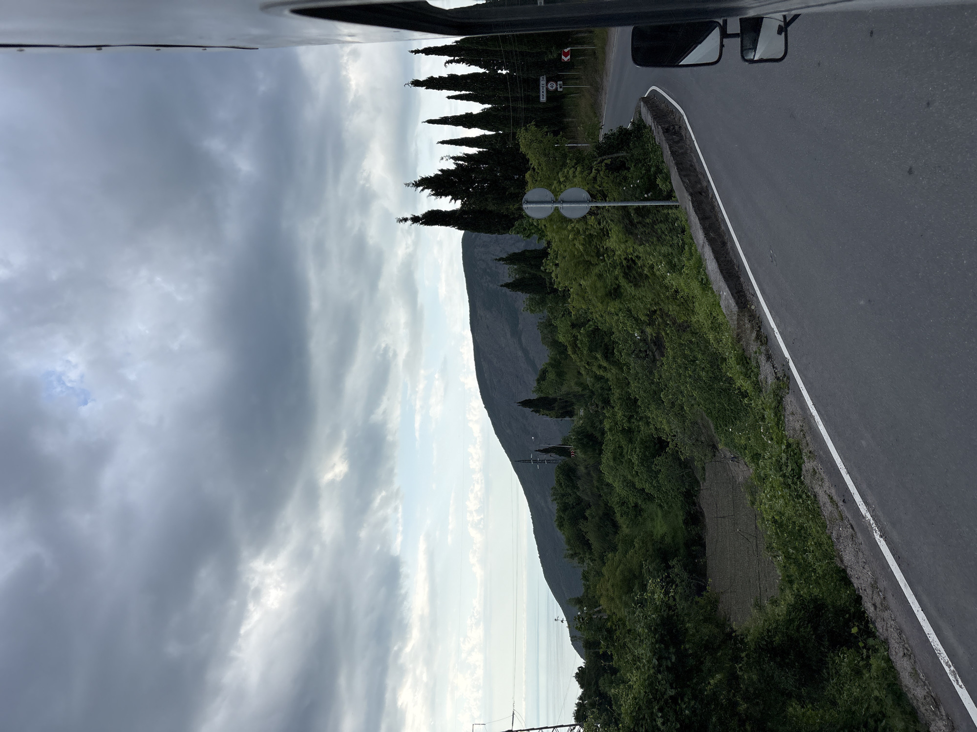 The image shows a view from a vehicle driving on a road. To the left, a cloudy sky dominates. On the right side of the road, there's lush greenery and a mountain in the background. Road signs are visible along the road, and a white line marks the edge of the pavement. The specific location is Nizhnee Zaprudnoe, Ukraine.