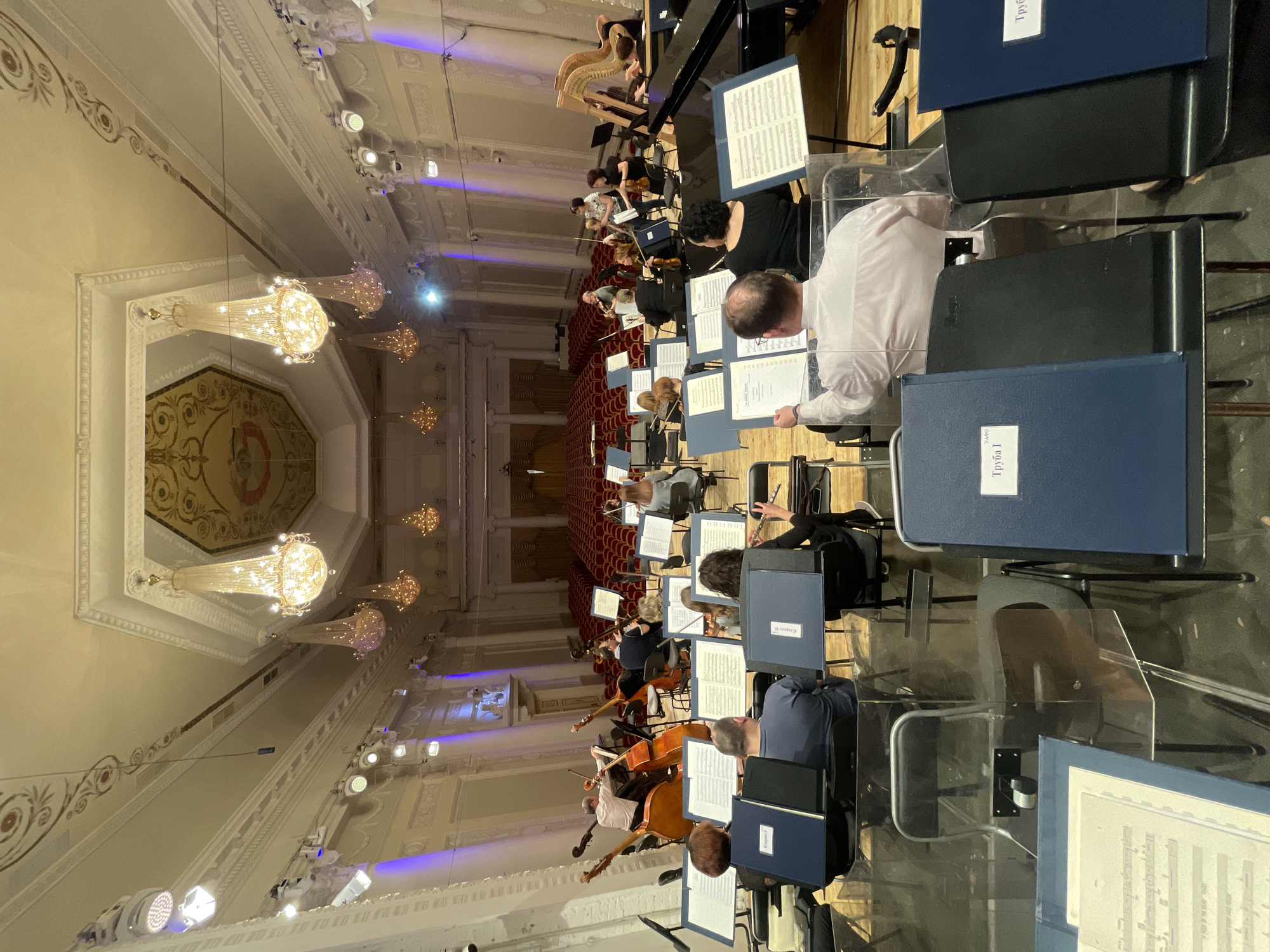 The image depicts an orchestra in performance inside a grand hall. Numerous musicians are seated in rows, equipped with music stands and sheet music. Instruments visible include violins, cellos, a harp, and possibly a piano in the background. The hall features ornate ceilings with chandeliers. From the text visible on the music stands in the foreground, it can be inferred that the orchestra is located at the Sverdlovsk State Academic Philharmonic, in Yekaterinburg, Russia. Some of the musicians are looking at their sheet music; others are actively playing their instruments. Audience seating is visible at the back of the hall and appears to be unoccupied.
