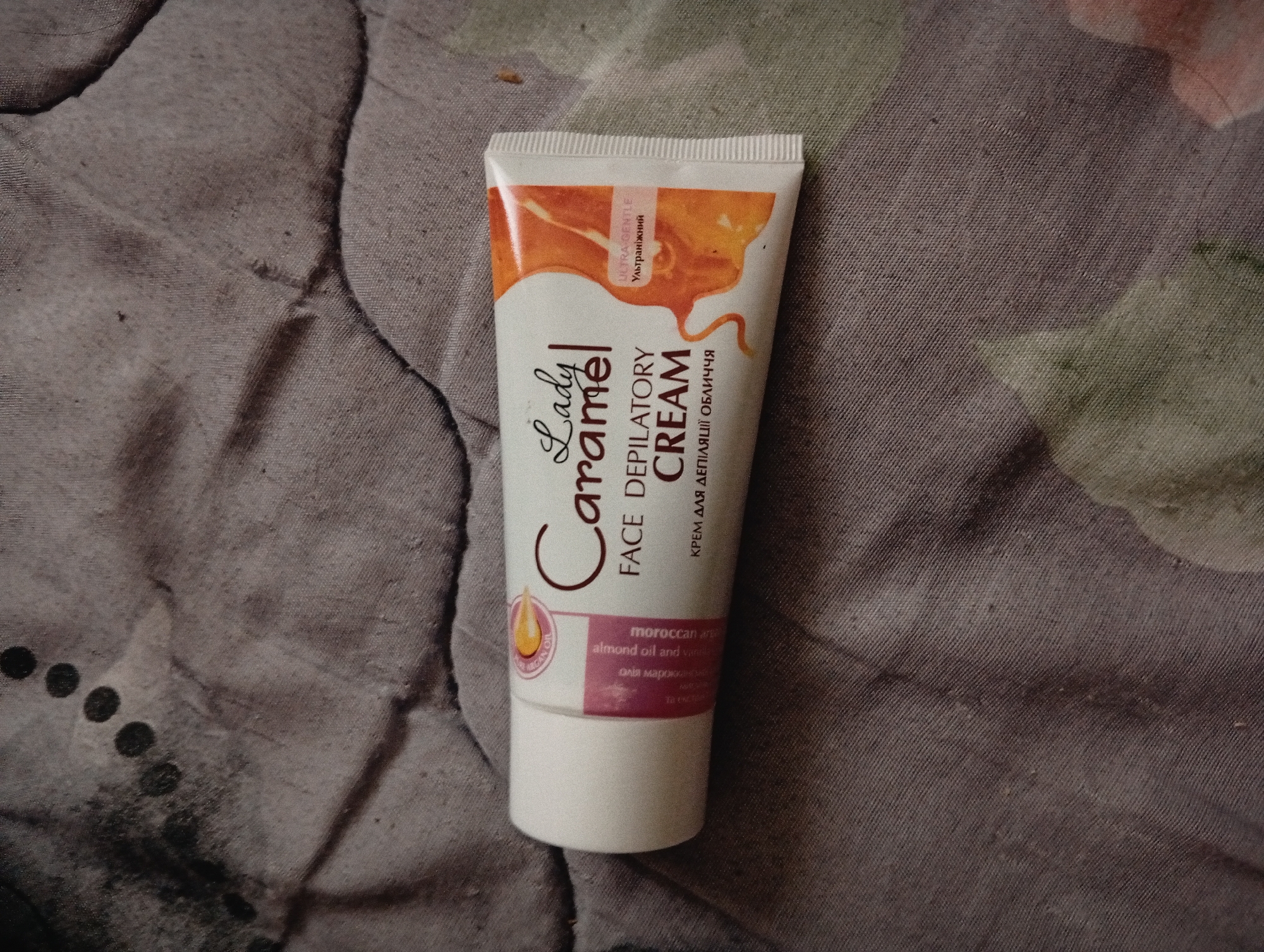 The image shows a tube of "Lady Caramel" face depilatory cream lying on a patterned fabric surface. The cream is in a white tube with caramel-colored accents and text in both English and Ukrainian. The text on the tube includes "Lady Caramel," "Face Depilatory Cream," "Ultra Gentle," "Moroccan Argan Almond Oil and Vanilla" and their Ukrainian translations. The background fabric has a muted color palette, showing various shades of brown, purple, and green, with visible texture. The details related to location, namely the city of Poltava, Ukraine, are not discernible from the image.