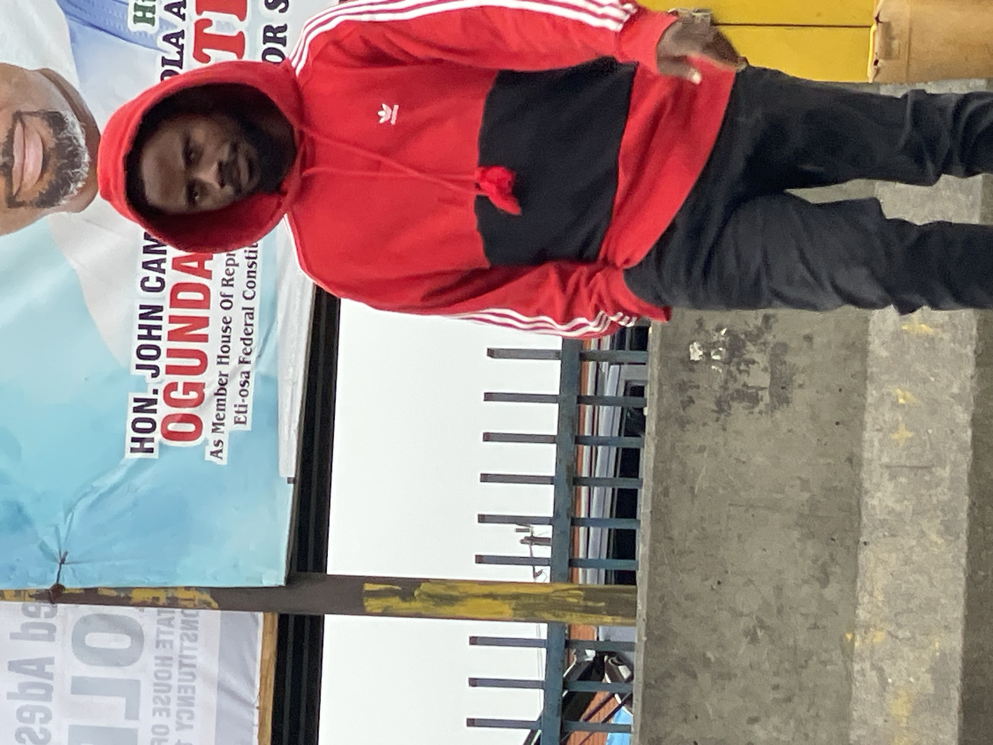 A medium shot photograph features a dark-skinned man wearing a red and black Adidas hoodie, black pants, and sneakers. He is positioned in front of political campaign posters. One banner features a headshot of the candidate, HON. JOHN CAM OGUNDA, for the House of Representatives, Eti-osa Federal Constituency. The man's posture is slightly tilted to the left. He is facing the camera, and his hand is lifted towards his side.
The scene is in an urban environment, likely in Ado, Nigeria, as indicated by the political campaign posters, architectural elements, and the person. A partially visible fence and what looks like a street structure are in the background. The concrete of the walkway is also visible.