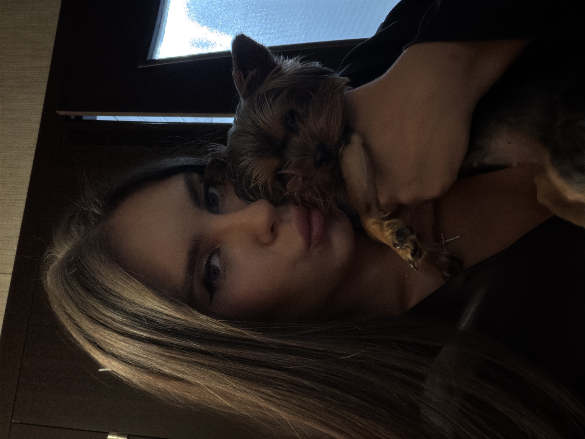 A woman with long, light-brown hair is holding a small, dark-brown dog close to her face. The background includes a window with partially visible daylight. The location is assumed to be indoors. The woman is wearing a dark-colored top.