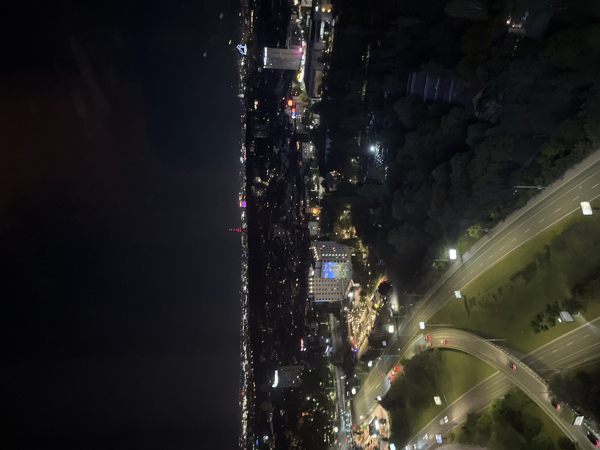 This high-angle, nighttime photograph captures an expansive view of a city, identified as Sofia, Bulgaria. The scene is dominated by a complex network of illuminated roads, countless artificial lights from buildings, and a vast, dark body of water to the left, possibly a sea or large lake, reflecting faint distant lights.
The urban landscape stretches horizontally, with city lights extending far into the horizon along the waterfront. In the mid-ground, a multi-lane highway system with elevated overpasses and sweeping curves is prominent. Vehicles with visible headlights and red taillights are moving along these roads, appearing as short streaks of light due to the nighttime exposure. Bright streetlights uniformly illuminate the roadways.
The city is densely packed with a mix of buildings. Several high-rise structures stand out, along with numerous smaller, brightly lit buildings. A large building complex in the central right features a distinctive blue-lit section, possibly a facade or a large display. Dark, densely vegetated areas, likely parks or hillsides, are interspersed throughout the city, particularly to the right of the main road network. The overall impression is that of a bustling urban environment active after dark. No specific human activity is discernible, nor is any text visible. The time of day is clearly night.