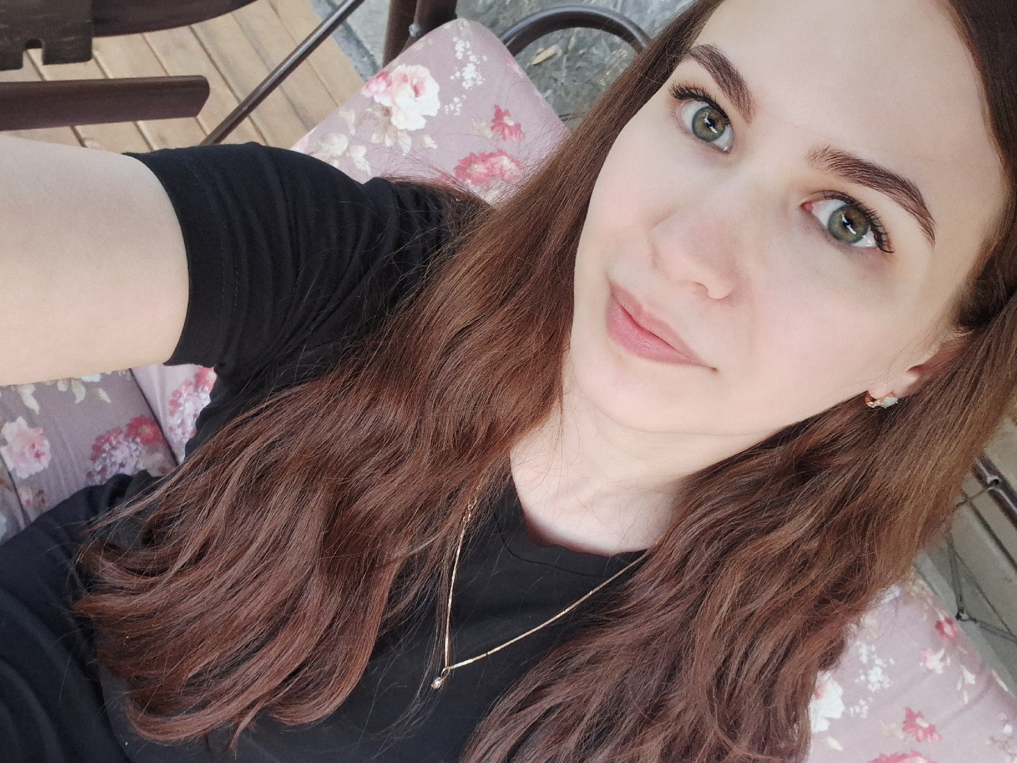 A selfie of a fair-skinned woman with long, auburn hair and green eyes is shown from a low-angle perspective. She is wearing a black T-shirt and a delicate gold necklace with a small pendant. Her earrings are visible. The background consists of a floral-patterned cushion and what appears to be a wooden chair or swing. There is no information to confirm the location in the city of Malyi Bukor, Russia.