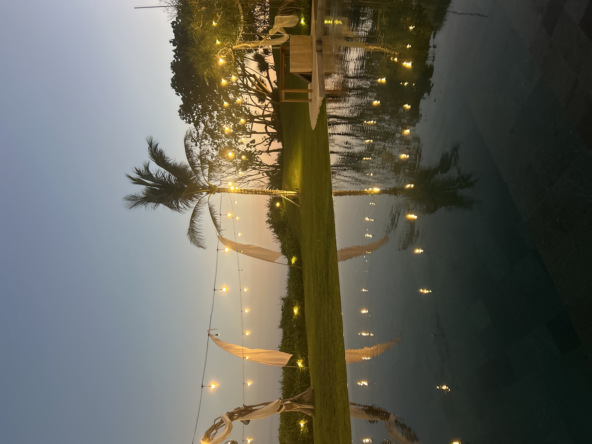 The image depicts an outdoor scene at dusk, likely a resort or a private villa, featuring a swimming pool in the foreground. String lights with warm yellow bulbs are strung across the scene, illuminating the lawn and reflecting on the still surface of the pool. A palm tree stands prominently on the right side of the frame, with other lush vegetation and trees in the background. Several white flags or banners are also visible, attached to wooden poles on the left side of the lawn. The sky is a soft gradient of blue and orange, indicating the transition from day to night. The reflection in the pool clearly shows the string lights and the surrounding trees and palm tree, creating a symmetrical effect. There is no indication of the specific location within the city of Kediri, Indonesia. No people or actions are depicted in the image.
